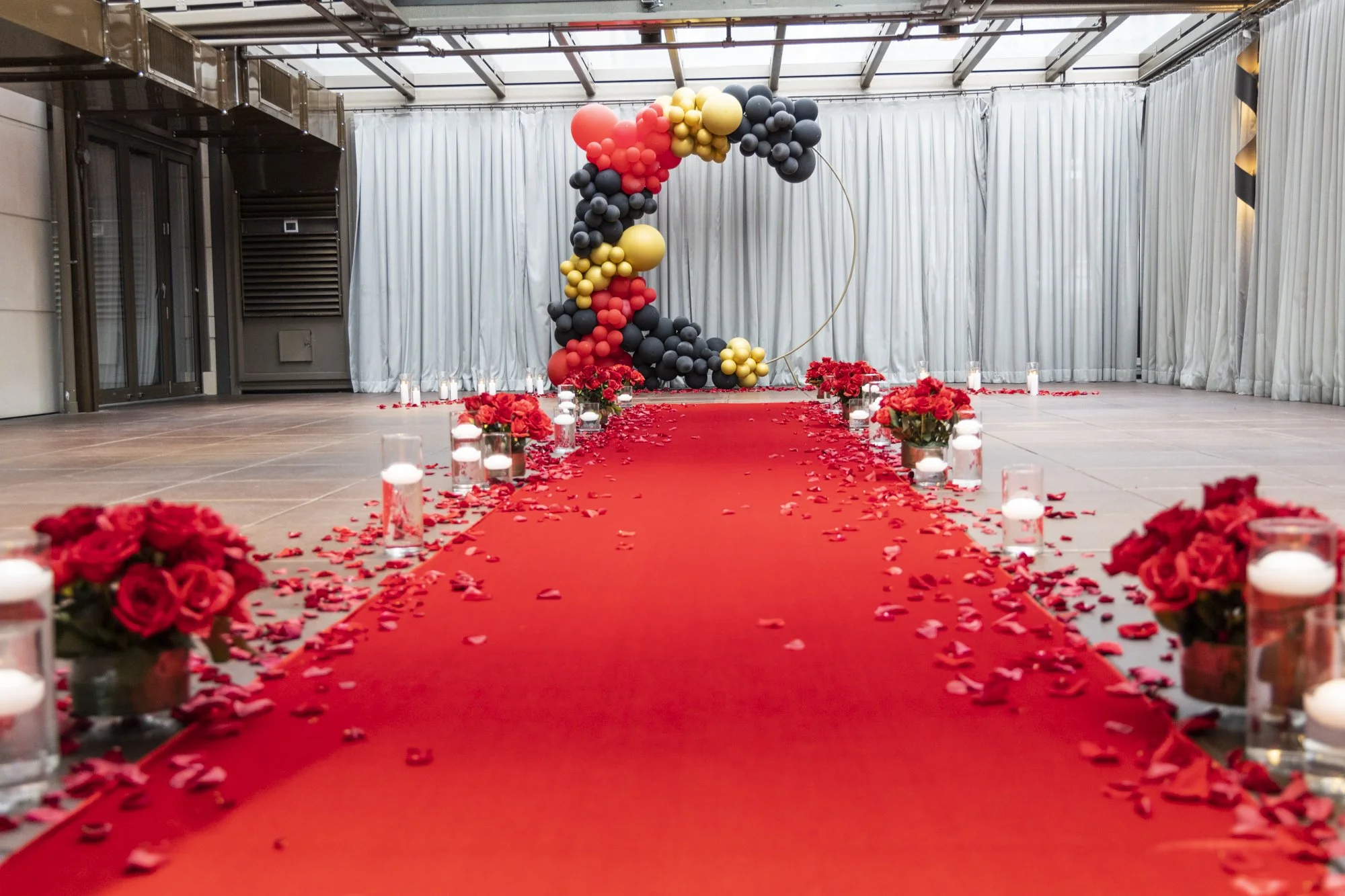A red carpet aisle decorated with scattered confetti, pink roses in vases, surrounded by small candles in glass holders, and an arch of black, red, and gold balloons at the end of the aisle.
