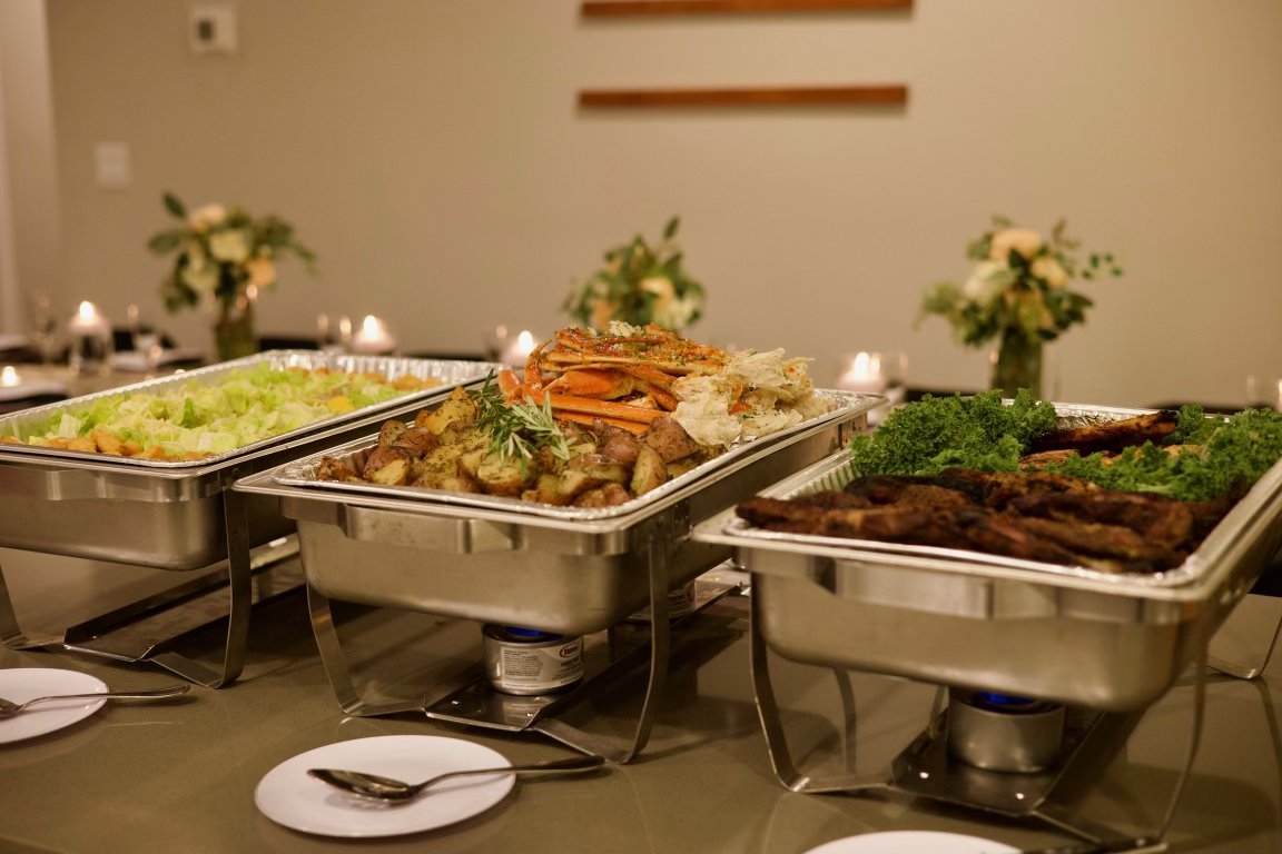 Buffet table with three chafing dishes containing salad, roasted vegetables, and grilled meat, set with plates and utensils in a dining room.