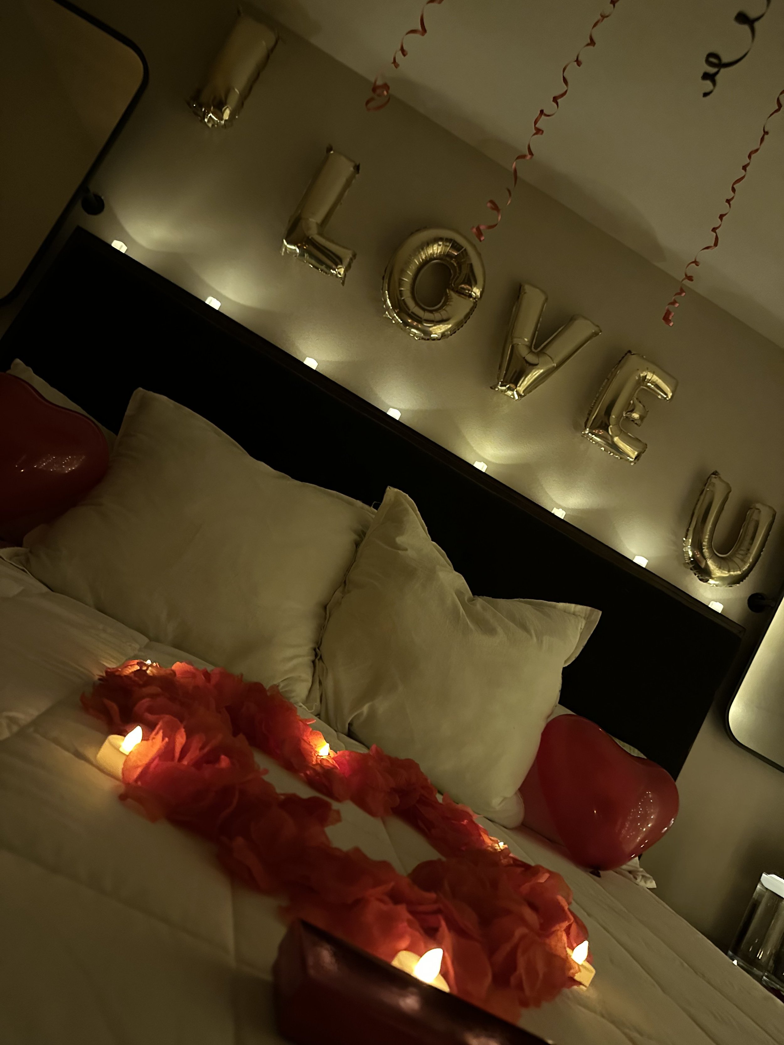 A decorated bedroom with gold letter balloons spelling 'LOVE' on the wall, pink heart-shaped balloons on the bed, and a heart-shaped arrangement of rose petals with small candles on the bed.