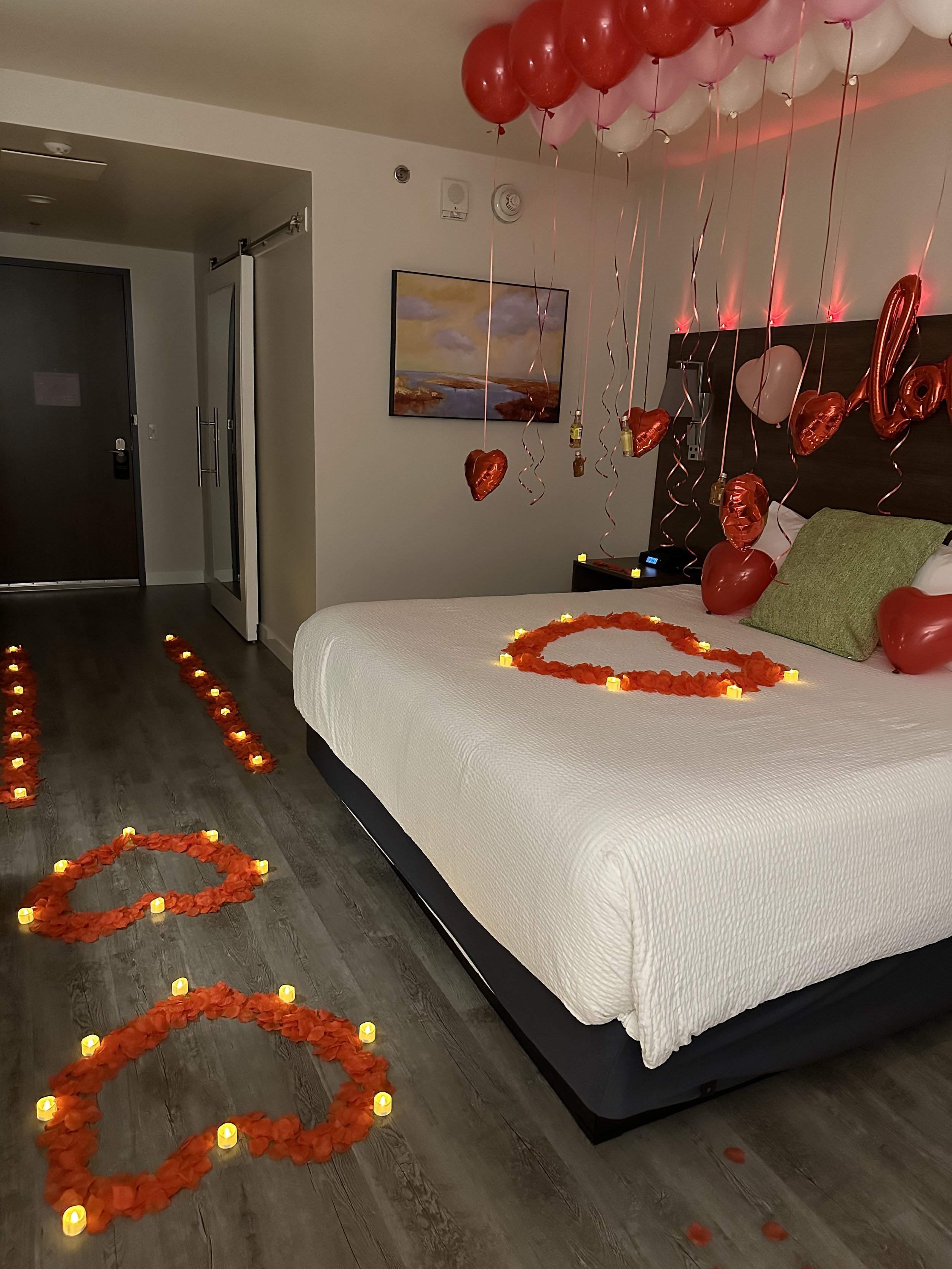 Hotel room decorated with red and pink balloons, heart-shaped balloons, rose petals, string lights, and candles forming heart shapes, indicating a romantic celebration.
