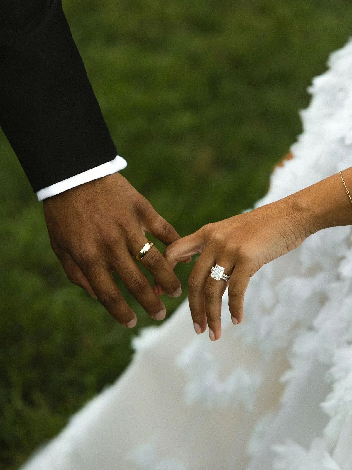 Close-up of a couple holding hands with wedding rings, one with a gold band, and the other with a diamond ring, against a grassy background with snow-like surface nearby.