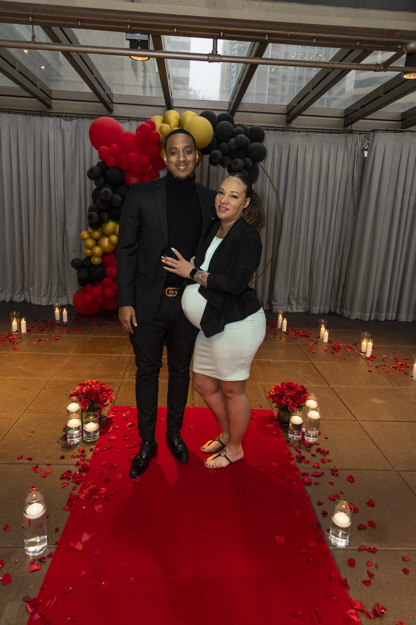 A couple stands together on a red carpet with rose petals and candles, during a romantic celebration, possibly a pregnancy announcement, in a decorated indoor venue with balloons and floral arrangements.