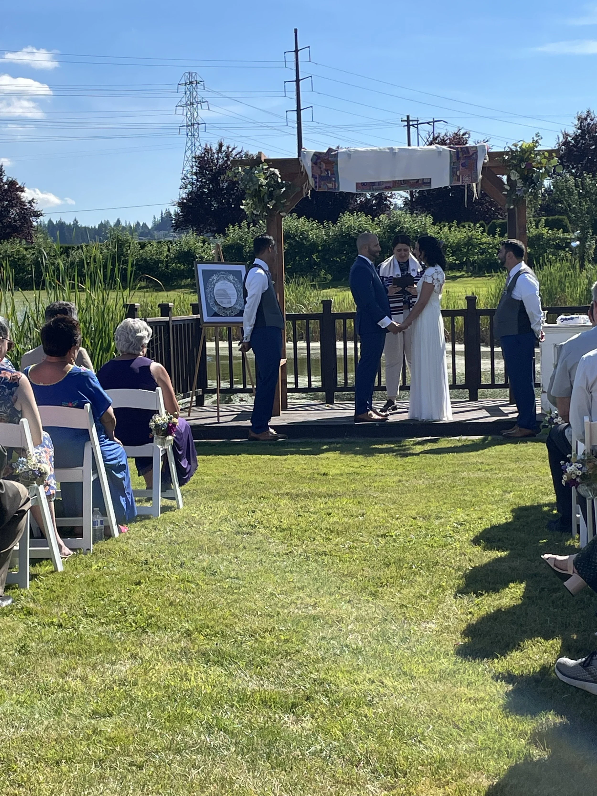 Couple getting married outdoors during a sunny day, standing under a wooden arch with officiant, guests seated on chairs, grassy field, and power lines in the background.
