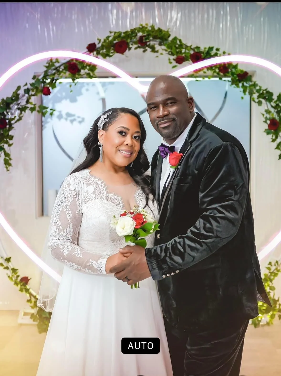 A newlywed couple in wedding attire, holding hands and smiling, standing in front of a heart-shaped floral decoration with roses.