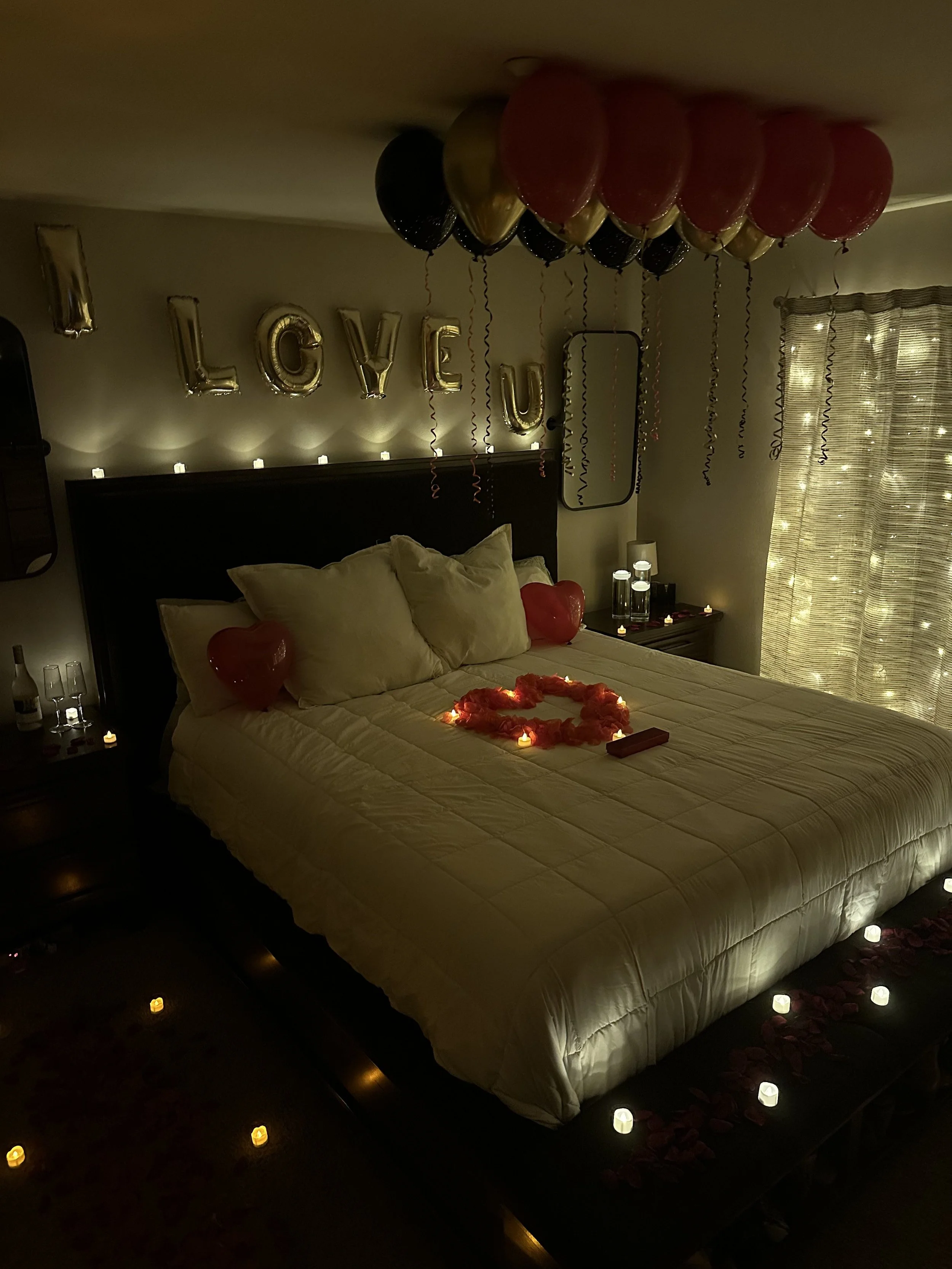 Decorated bedroom with balloons spelling 'LOVE U,' candles, heart-shaped balloons, a heart arrangement made of rose petals with candles inside it, and soft lighting.