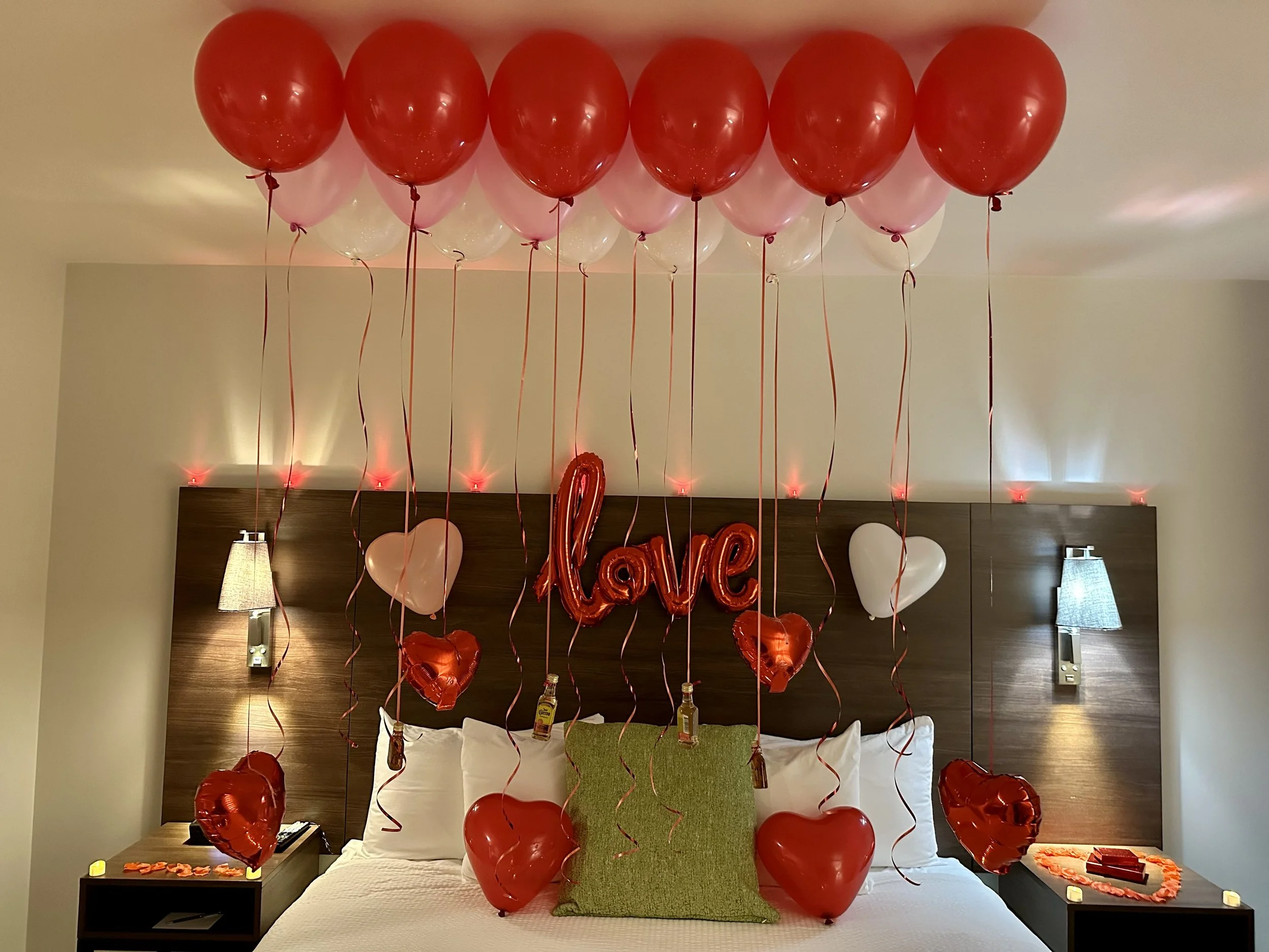 Decorated hotel room with red and pink balloons, heart-shaped balloons, 'love' balloon, and heart-shaped decor for a romantic occasion.