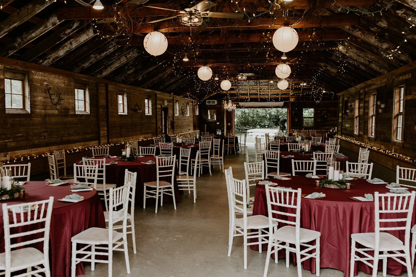 A rustic barn decorated for a celebration with string lights, paper lanterns, and tables with burgundy tablecloths and white chairs.