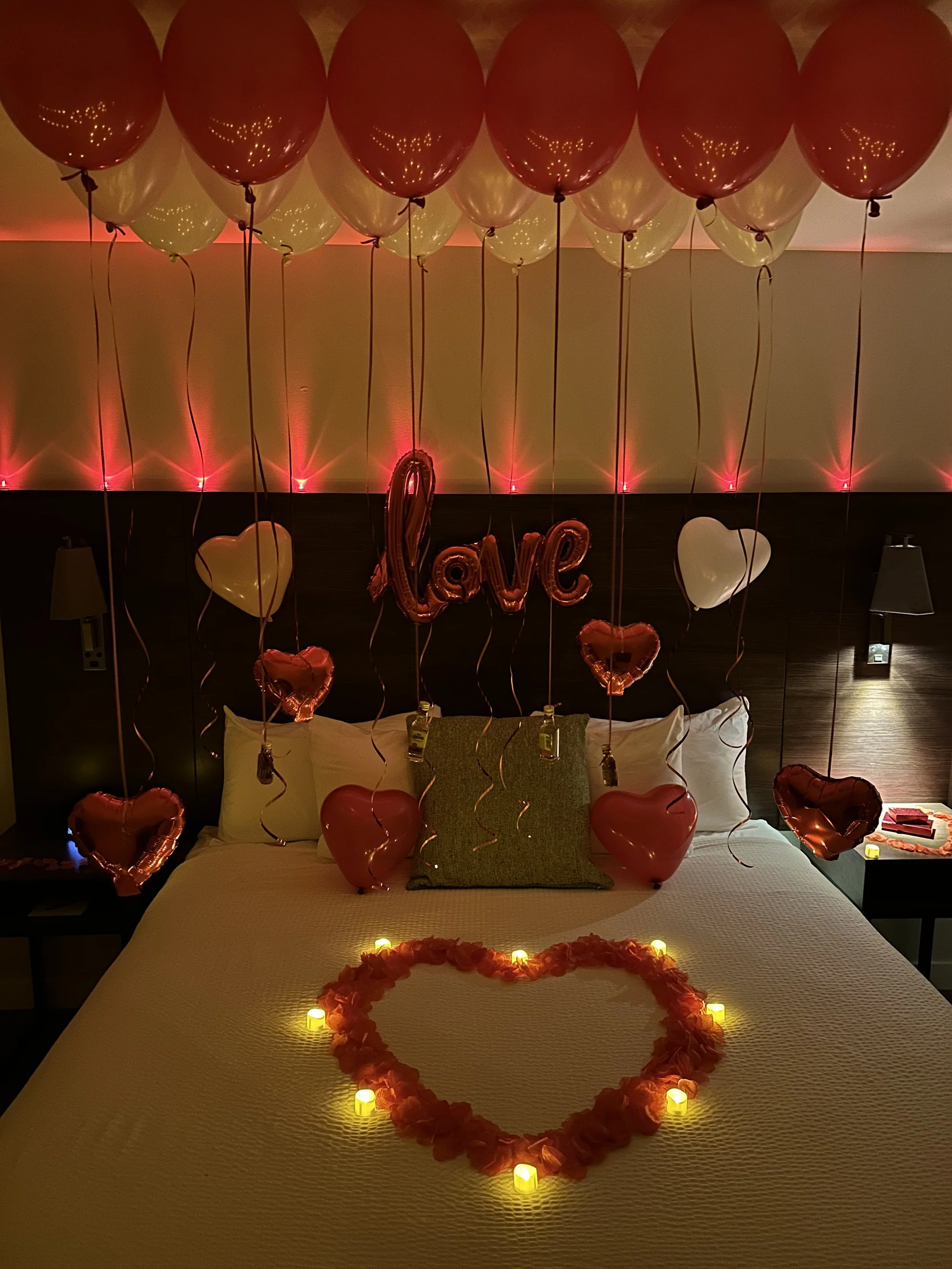 Decorated bedroom for Valentine's Day with red and white heart-shaped balloons, a heart made of rose petals with candles, and a 'love' balloon in the center.