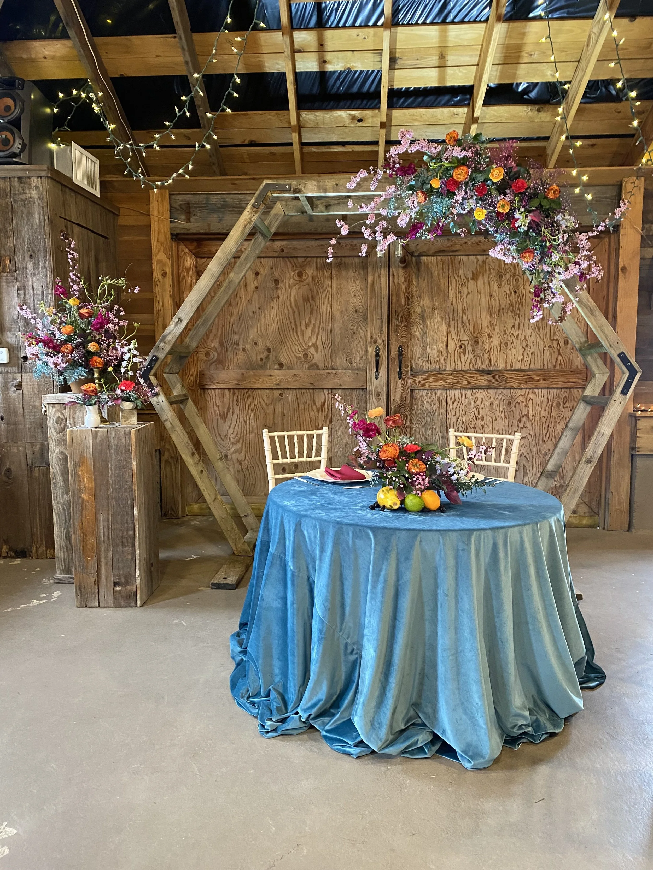 A rustic indoor setting featuring a round table with a blue satin tablecloth, decorated with floral centerpieces and fresh fruits. Behind the table is a wooden backdrop with a large, hexagonal wooden frame adorned with pink flowers. To the left is a 