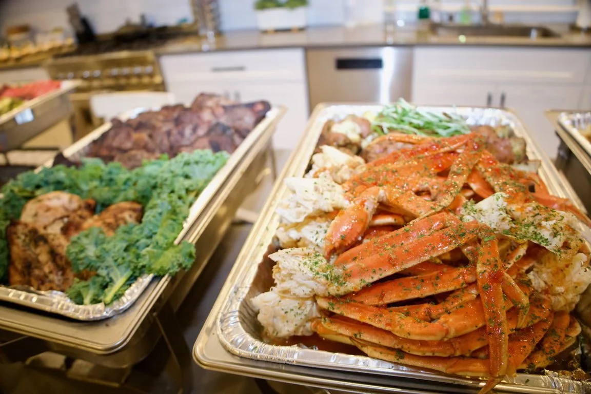 Trays of cooked seafood, vegetables, and meats on a buffet table inside a restaurant kitchen.