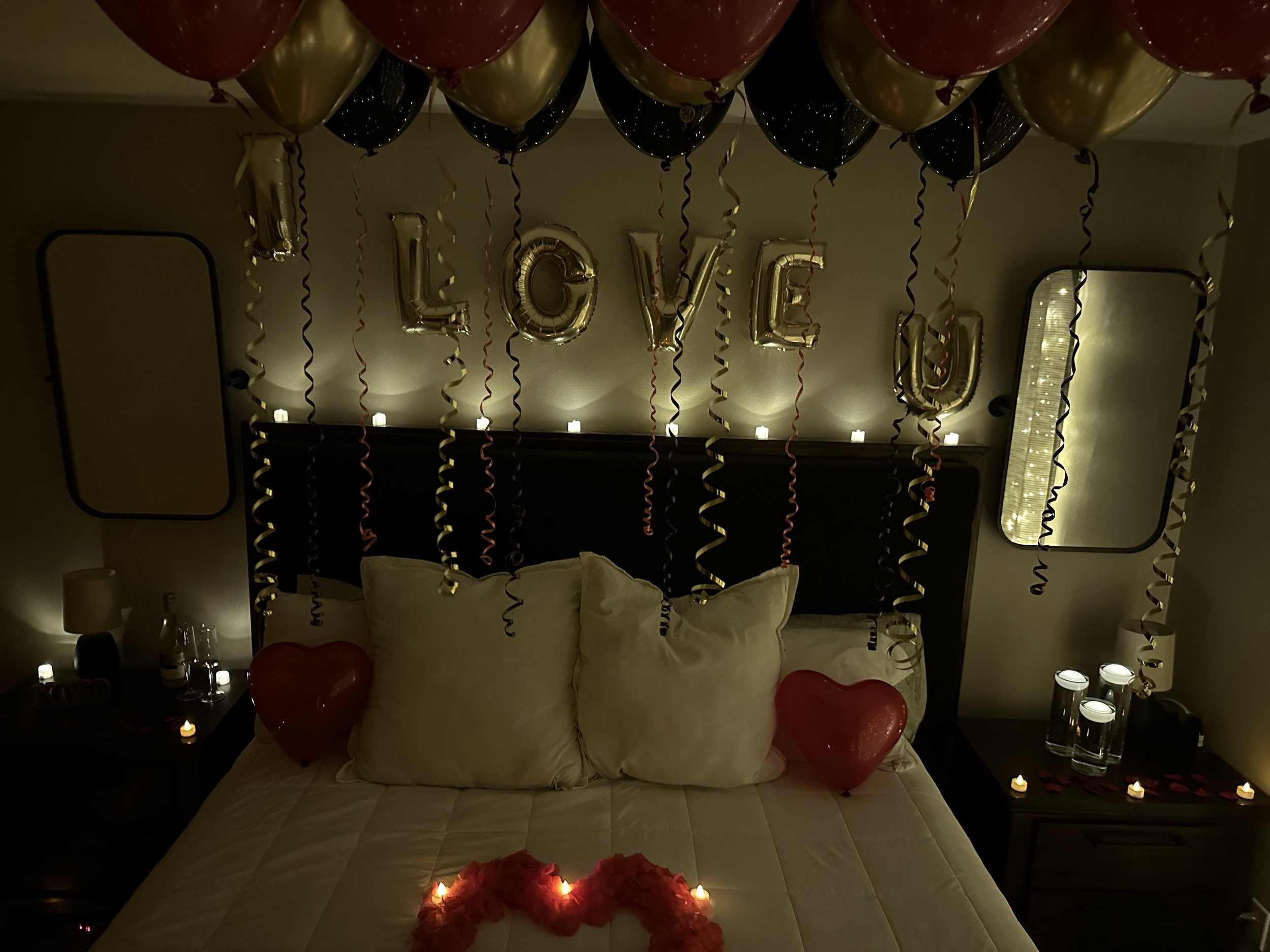 Decorated bedroom with love-themed balloons, candles, and the words 'LOVE U' in gold balloons on the wall, along with heart-shaped balloons and rose decorations on the bed.