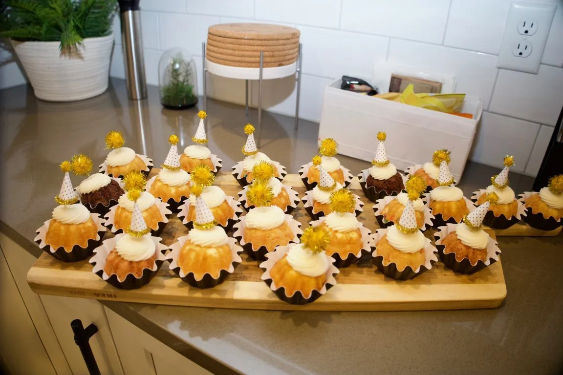 Cupcakes decorated with white frosting and yellow and white party hats with yellow pom-poms, arranged on a wooden serving board in a kitchen.