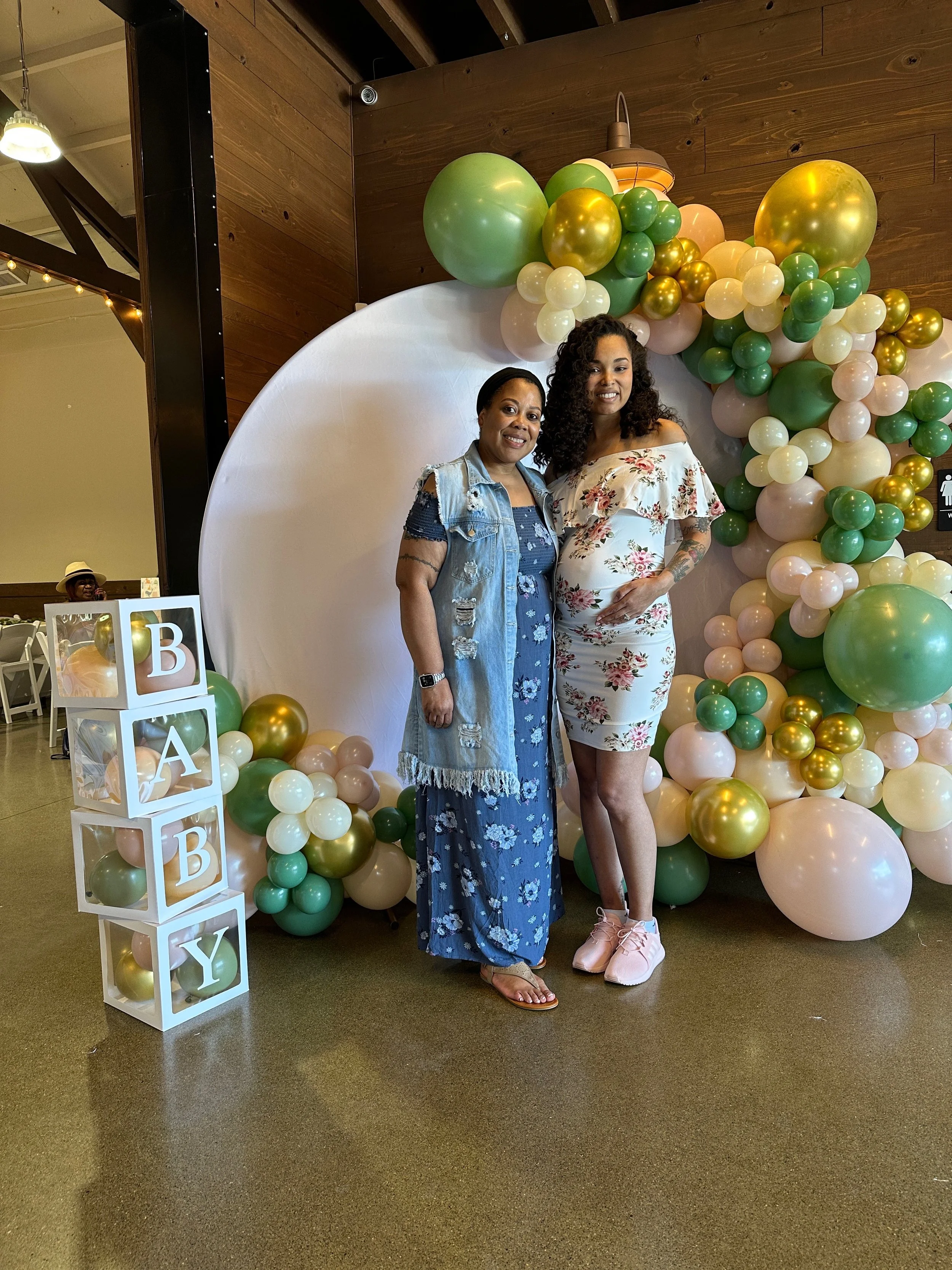 Two women standing in front of a large balloon arch with gold, green, cream, and pink balloons, beside a white cube display with the letters BABY, in an indoor venue with wooden walls.