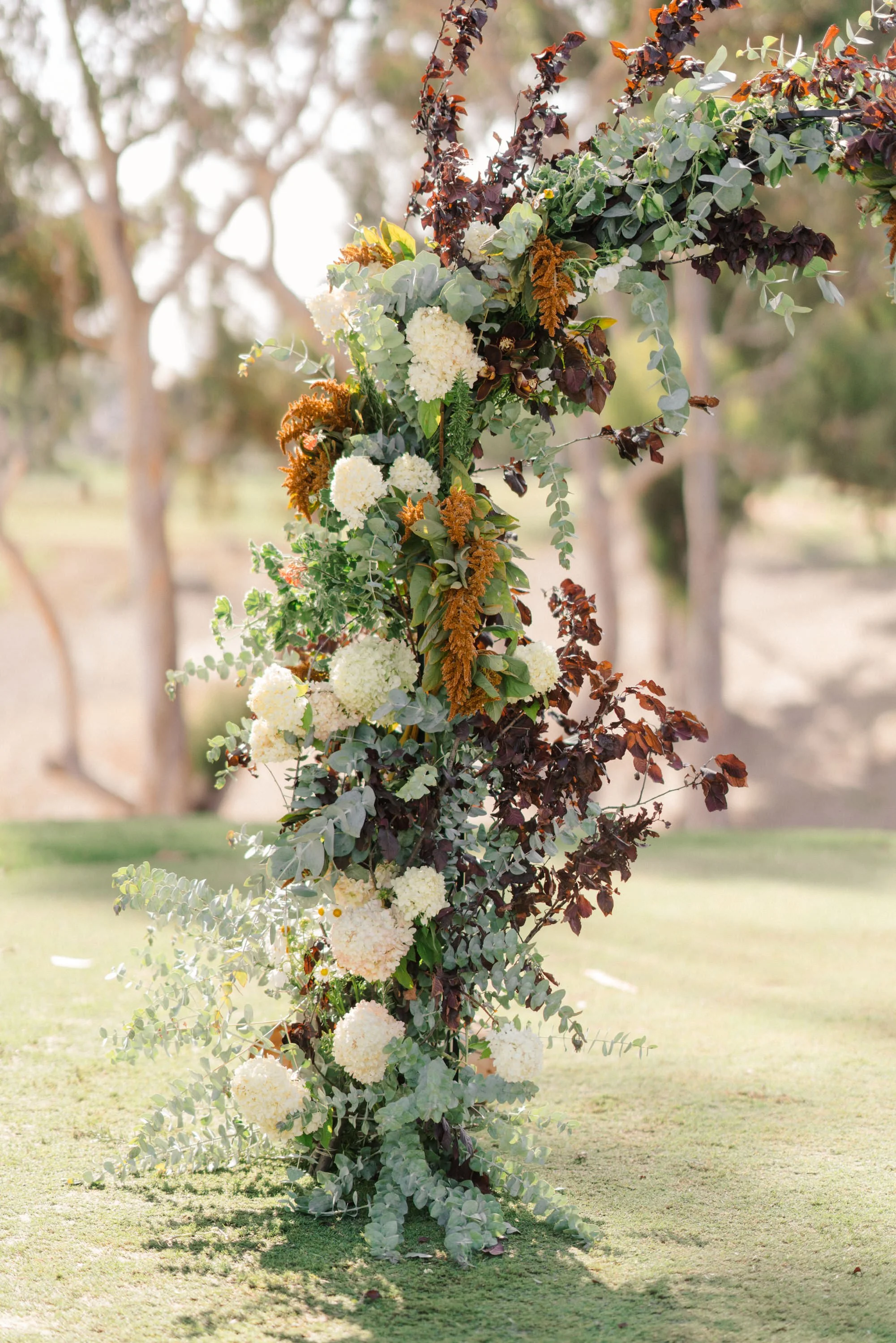A decorative floral arrangement with green and dark red and white flowers and leaves, set outdoors on a grassy area with trees in the background.