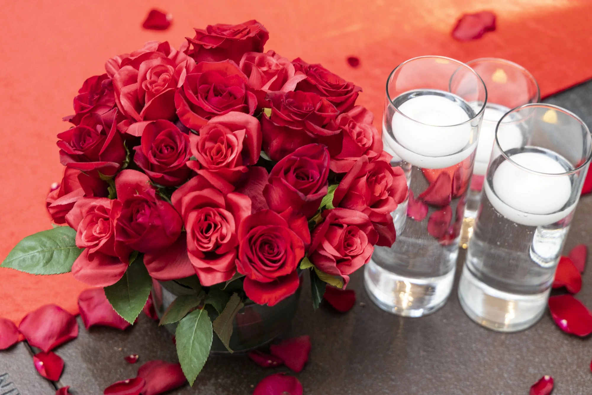 Bouquet of red roses with green leaves next to three glasses of water with floating rose petals on a table with scattered rose petals.