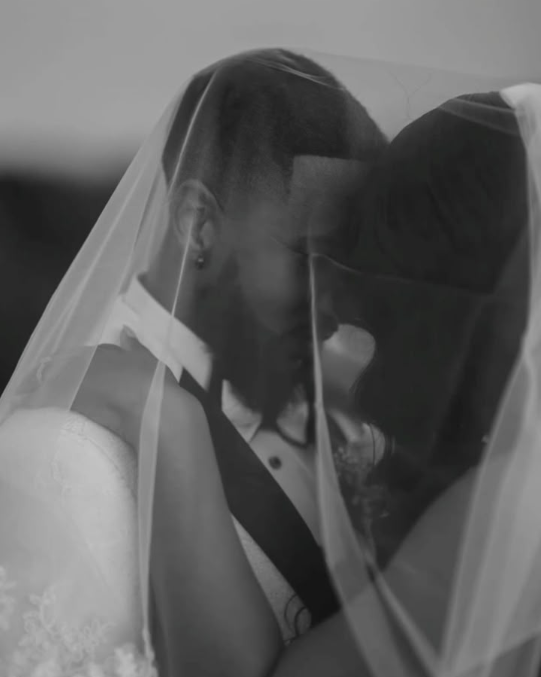 A black and white photo of a couple with foreheads touching, wearing a transparent face shield, during a wedding ceremony.