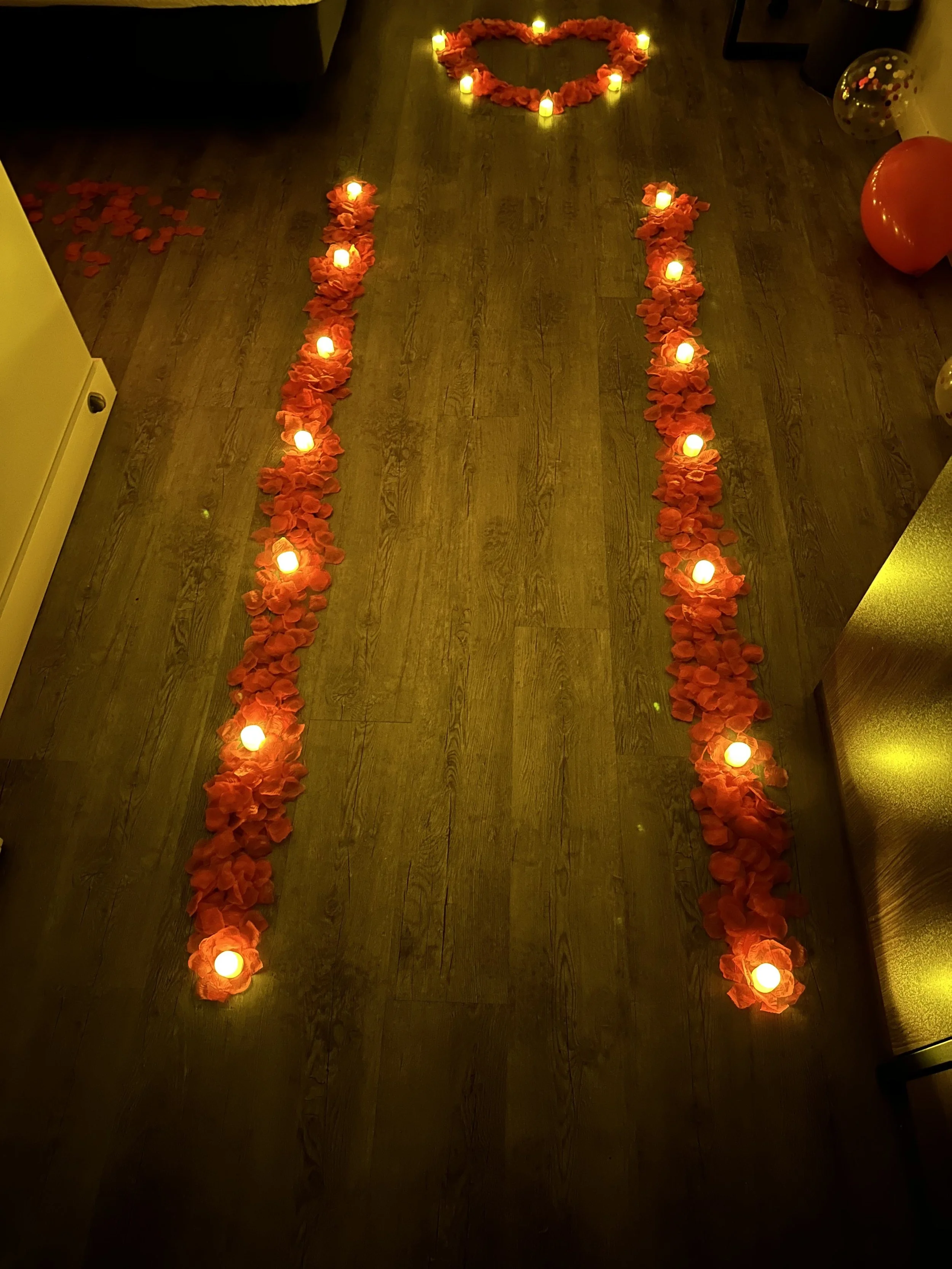 Candlelit heart shape arrangement with candles surrounded by flower petals at the top and a pathway lined with candles and flower petals on the floor.