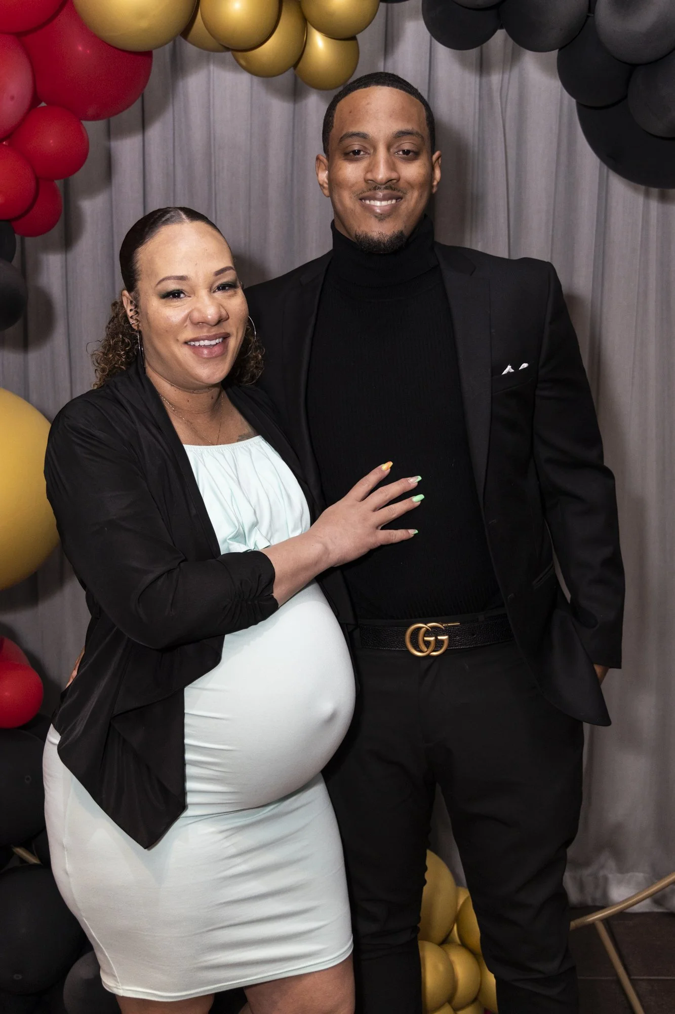 A pregnant woman in a white dress and black jacket standing next to a man in a black suit with a Gucci belt, posing for a photo in front of a balloon backdrop.