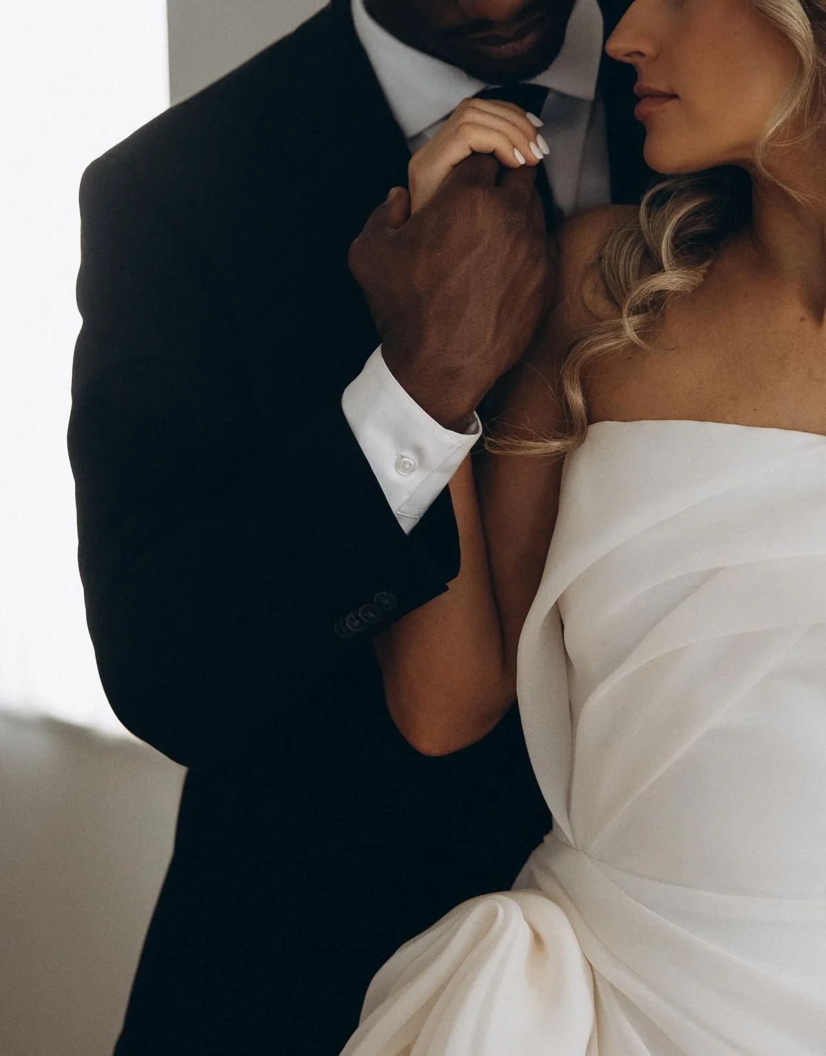 A close-up of a couple holding hands, with the man wearing a black suit and the woman in a white dress, possibly at a wedding or formal event.