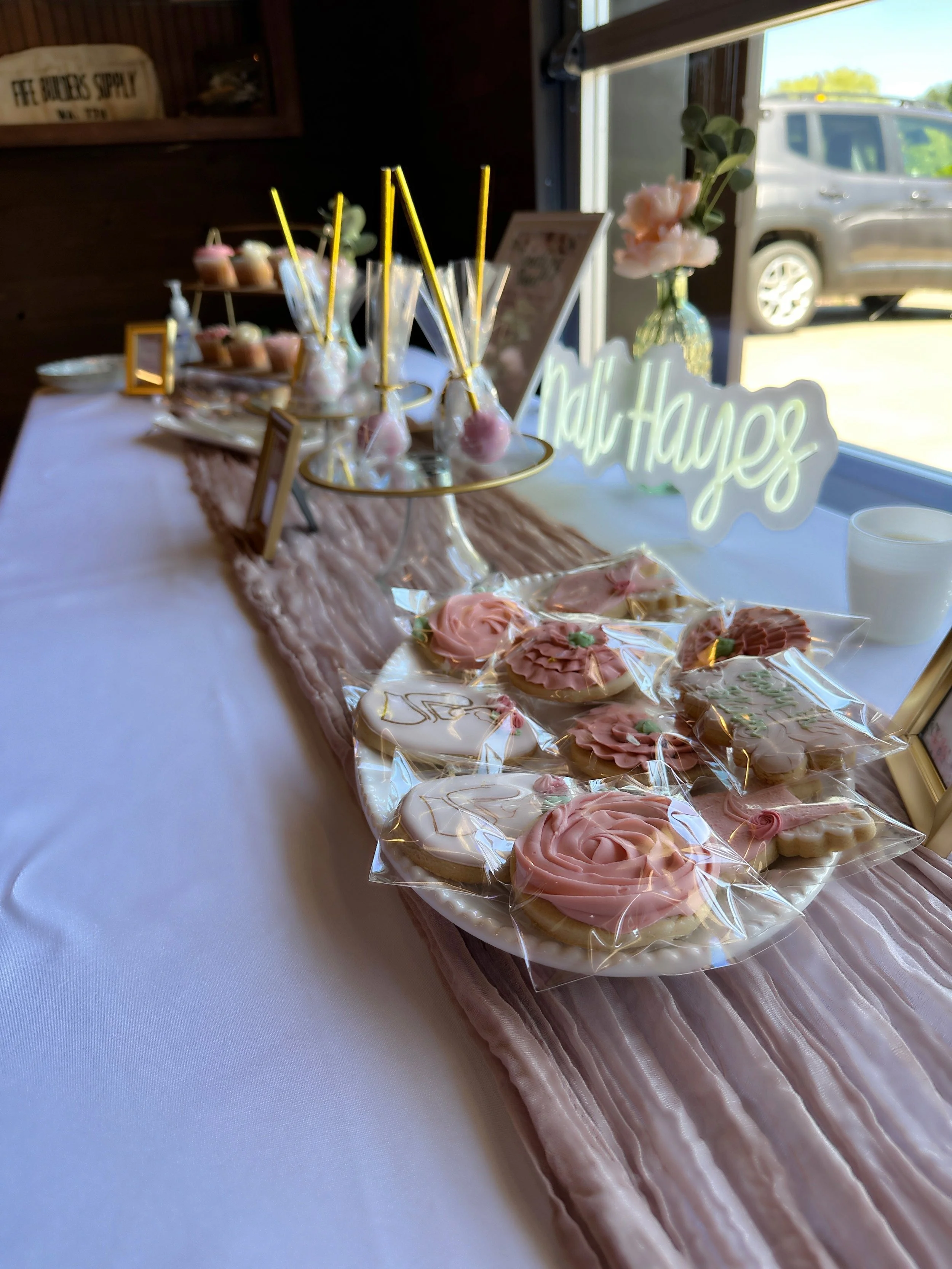 Decorative table with pink and white floral cookies, cake pops, and a "Hello Hays" sign near a window with a car outside.
