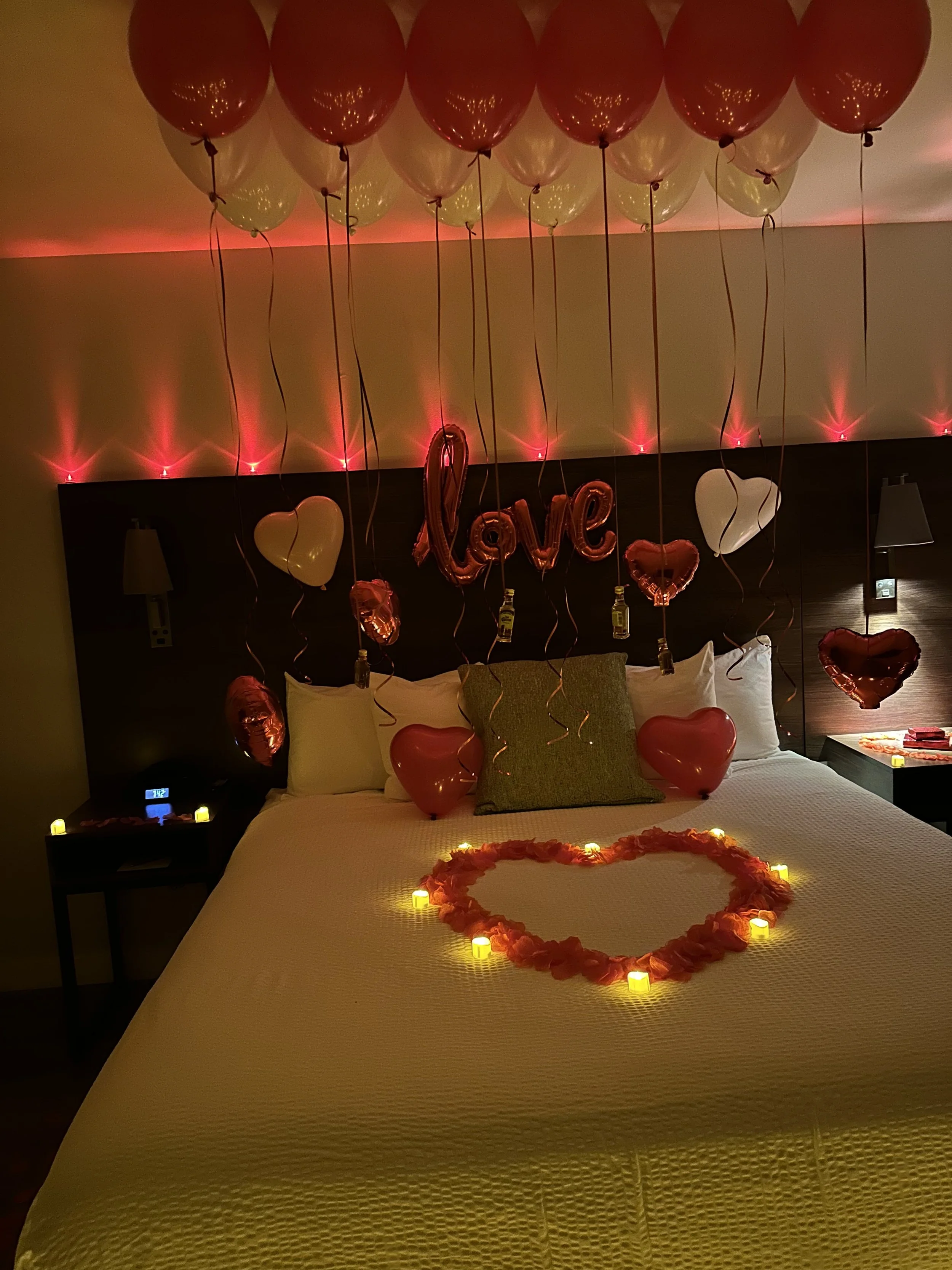 A bedroom decorated with balloons, candles, and romantic decor for Valentine's Day. There are heart-shaped balloons, a 'love' balloon, LED candles on the bed and its surroundings, and a heart-shaped arrangement of rose petals on the bed