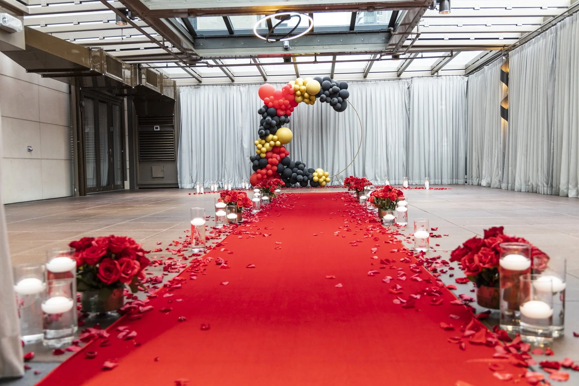Indoor wedding aisle decorated with red flower arrangements, candles, and rose petals, leading to a balloon arch with black, gold, red, and gray balloons, inside a glass-ceilinged venue.