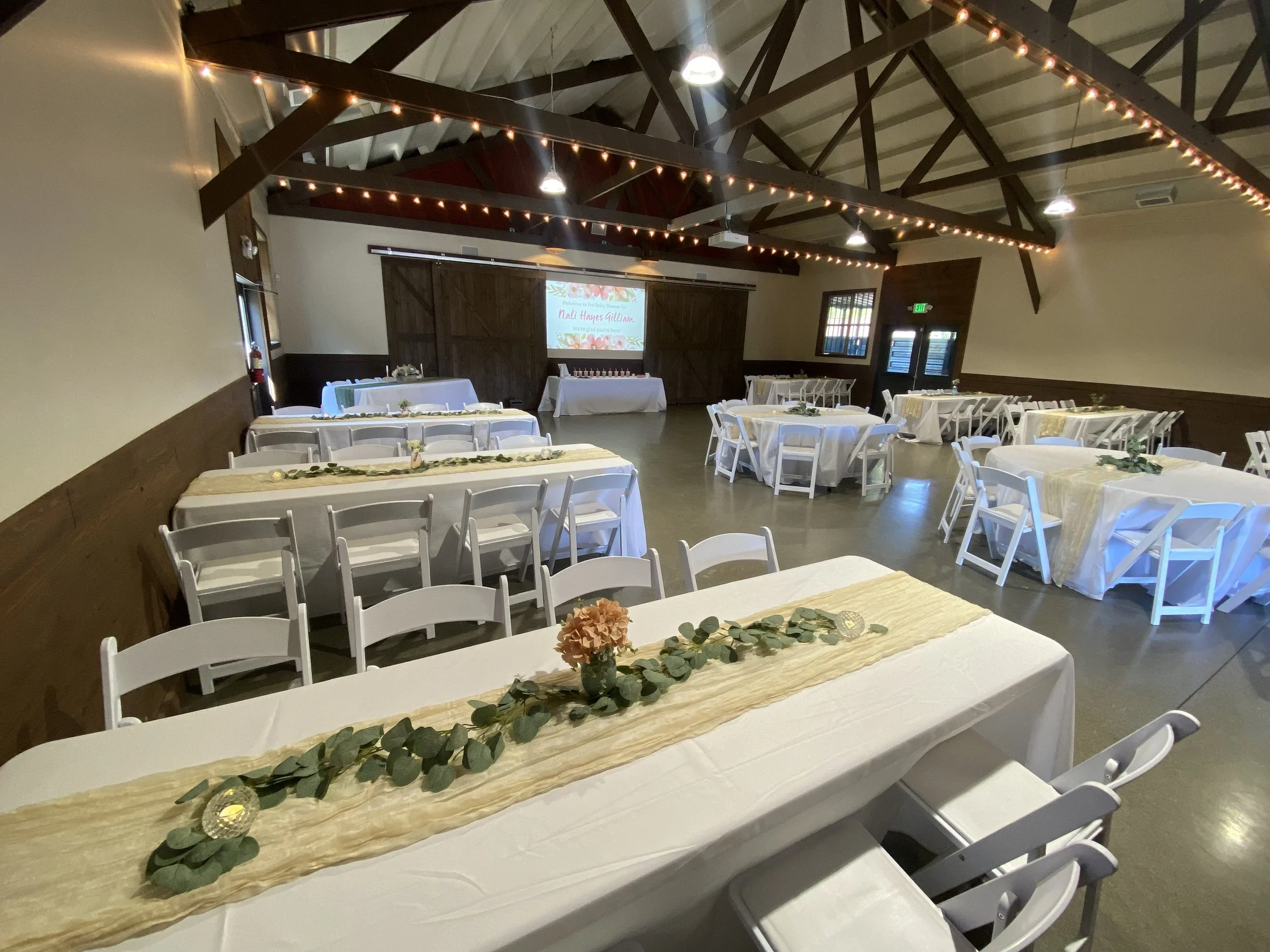 Event space decorated with long tables covered in white tablecloths and beige runner, adorned with greenery and candles. Wooden chairs surround the tables. The room has a high ceiling with exposed beams and hanging string lights. At the front, there 