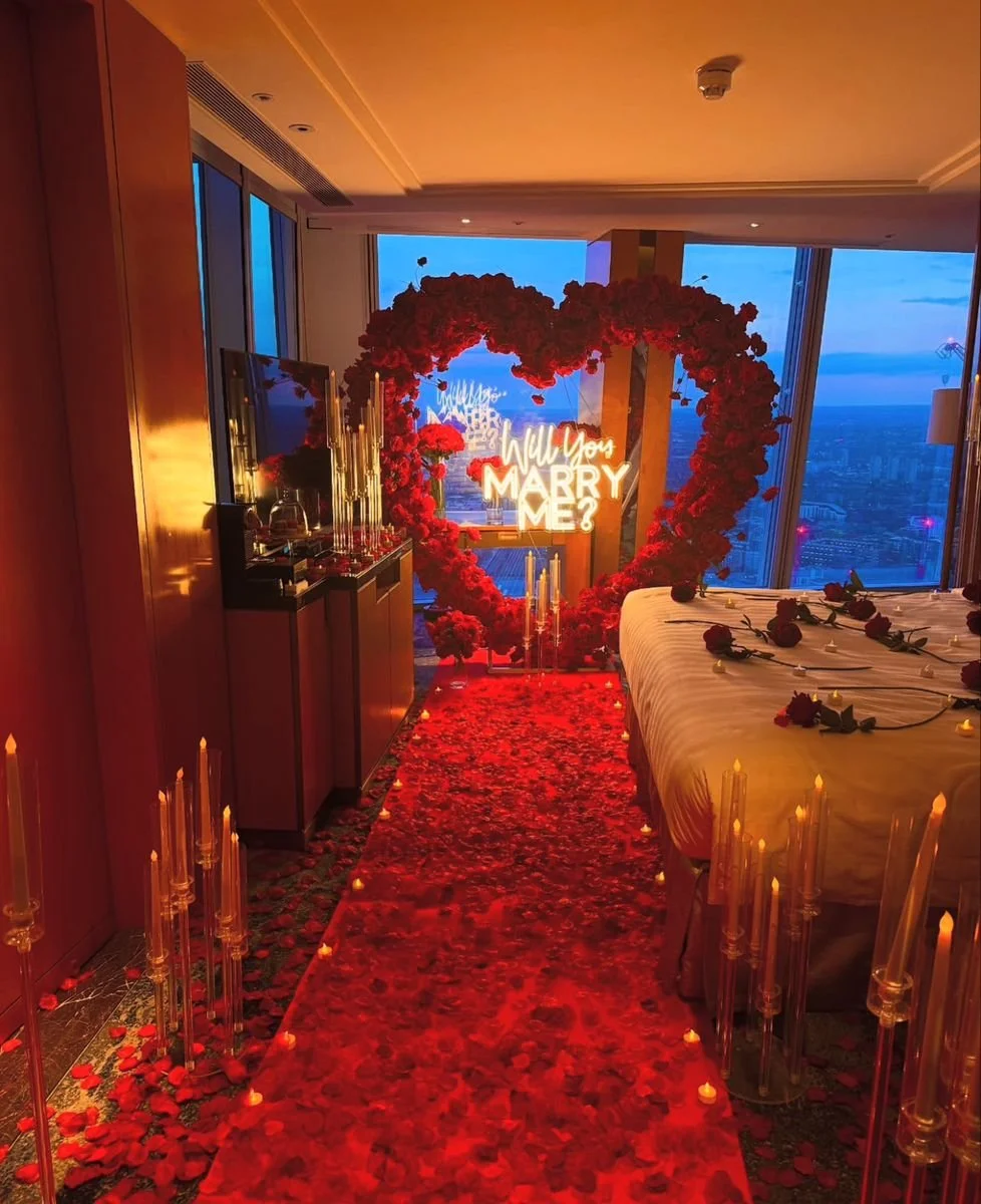 Elegant hotel room decorated for a marriage proposal with a large heart-shaped floral arch, red rose petals on the bed and floor, candles along the aisle, and a neon sign saying 'Will You Marry Me?' by the window with a city view at dusk.