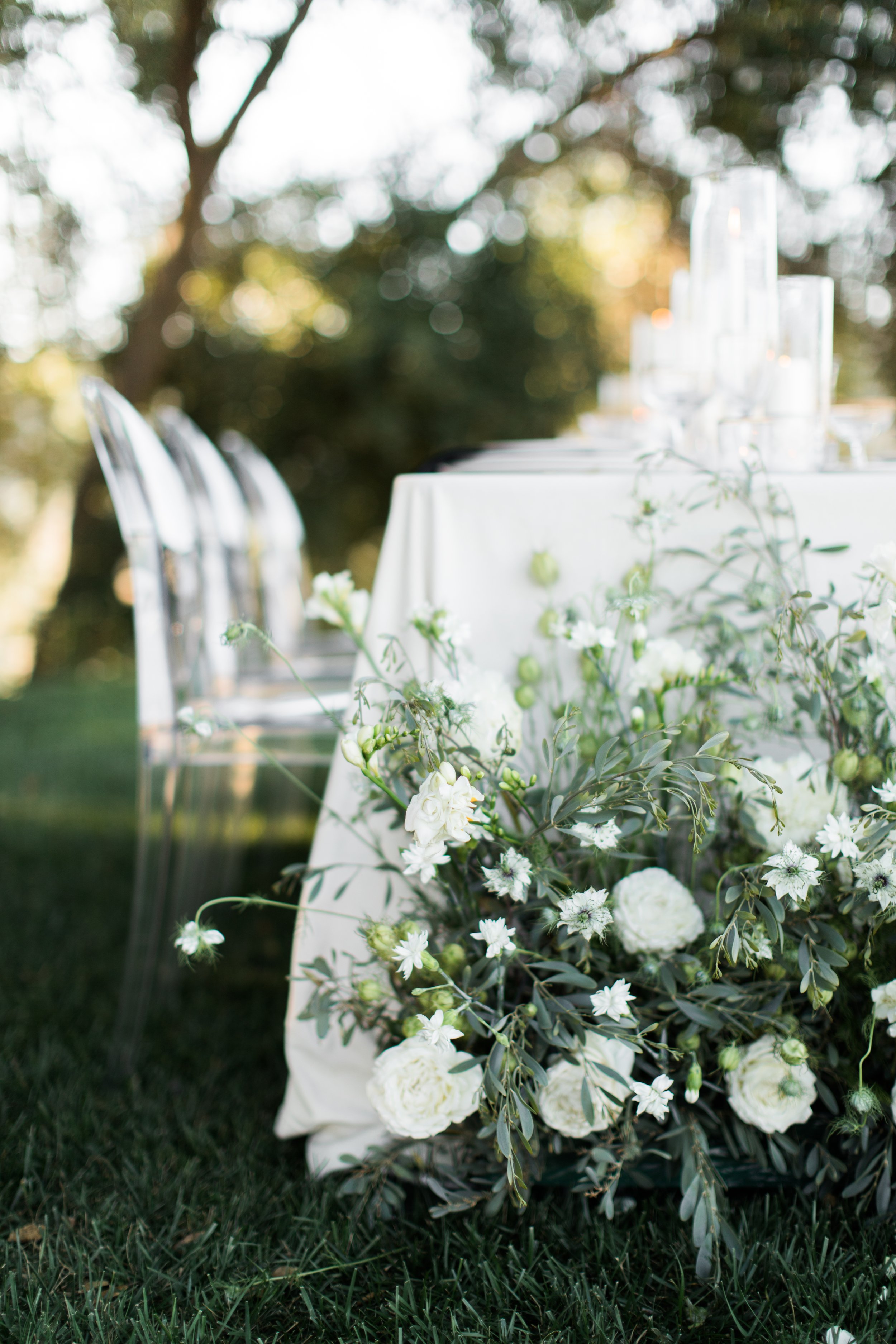 A table decorated with white flowers and greenery for an outdoor event, with glass candles and chairs in the background.