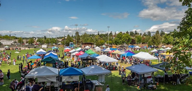 Outdoor fair with numerous vendor tents and crowds on a field, under a partly cloudy sky.