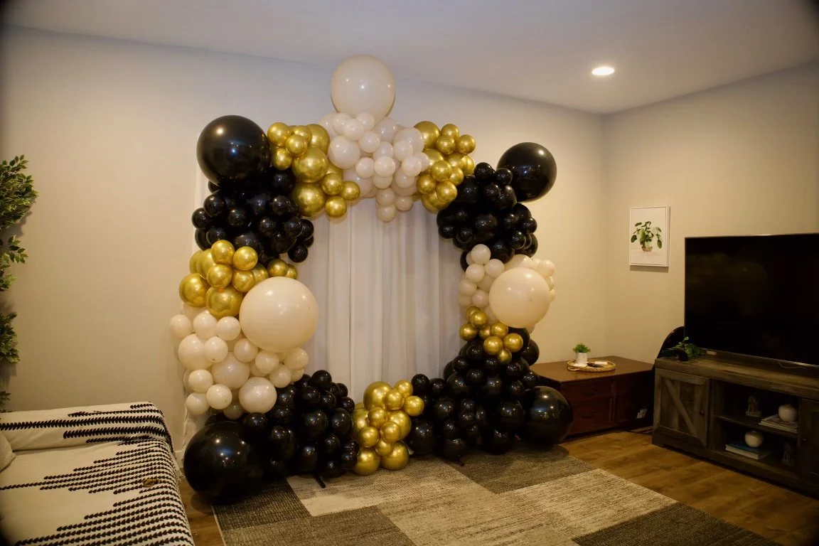 Balloon arch made of black, white, and gold balloons around a window or doorway in a living room.
