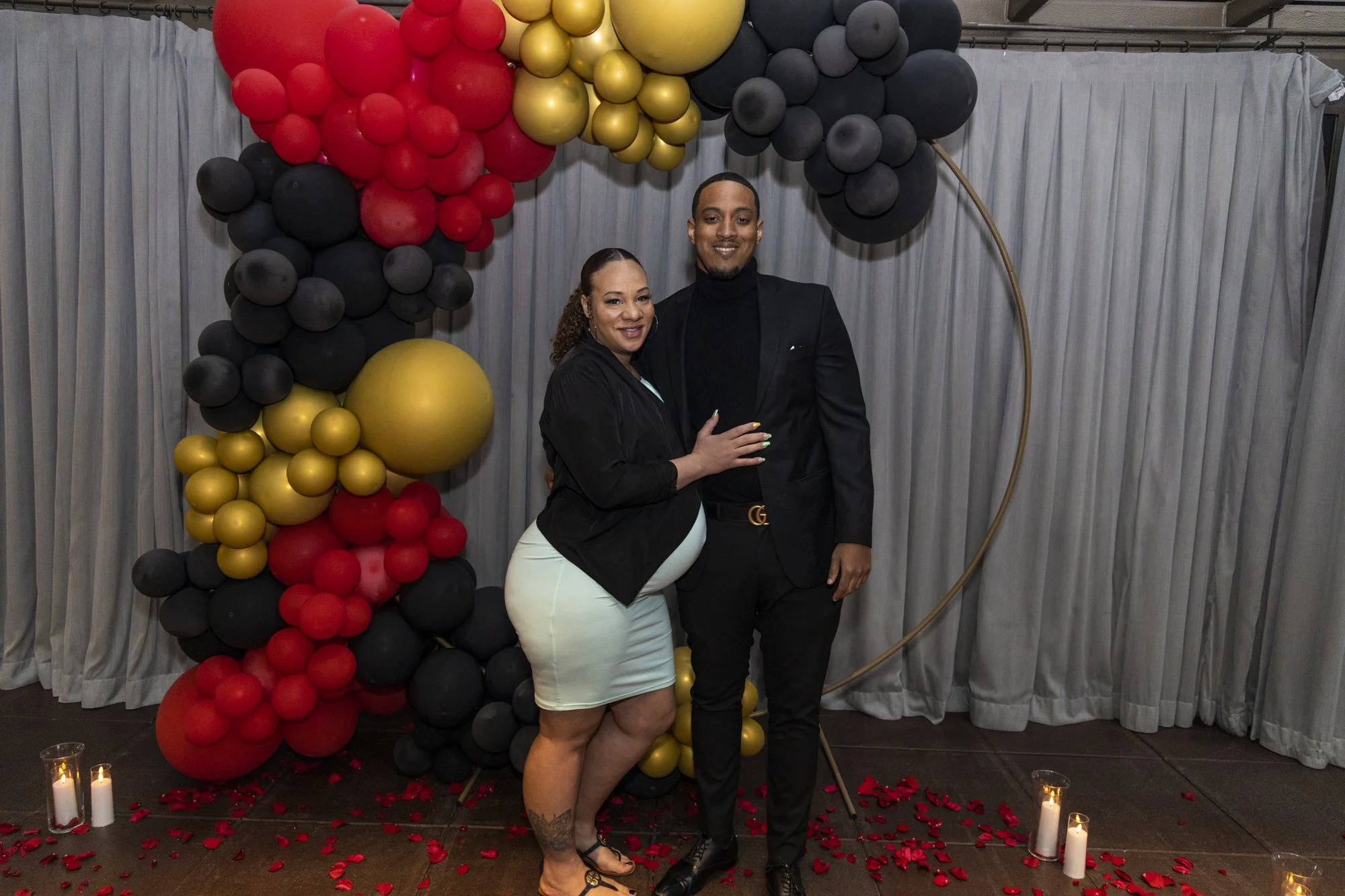 A couple stands together smiling in front of a balloon arch with black, red, and gold balloons, and candles on the floor with red rose petals scattered around.