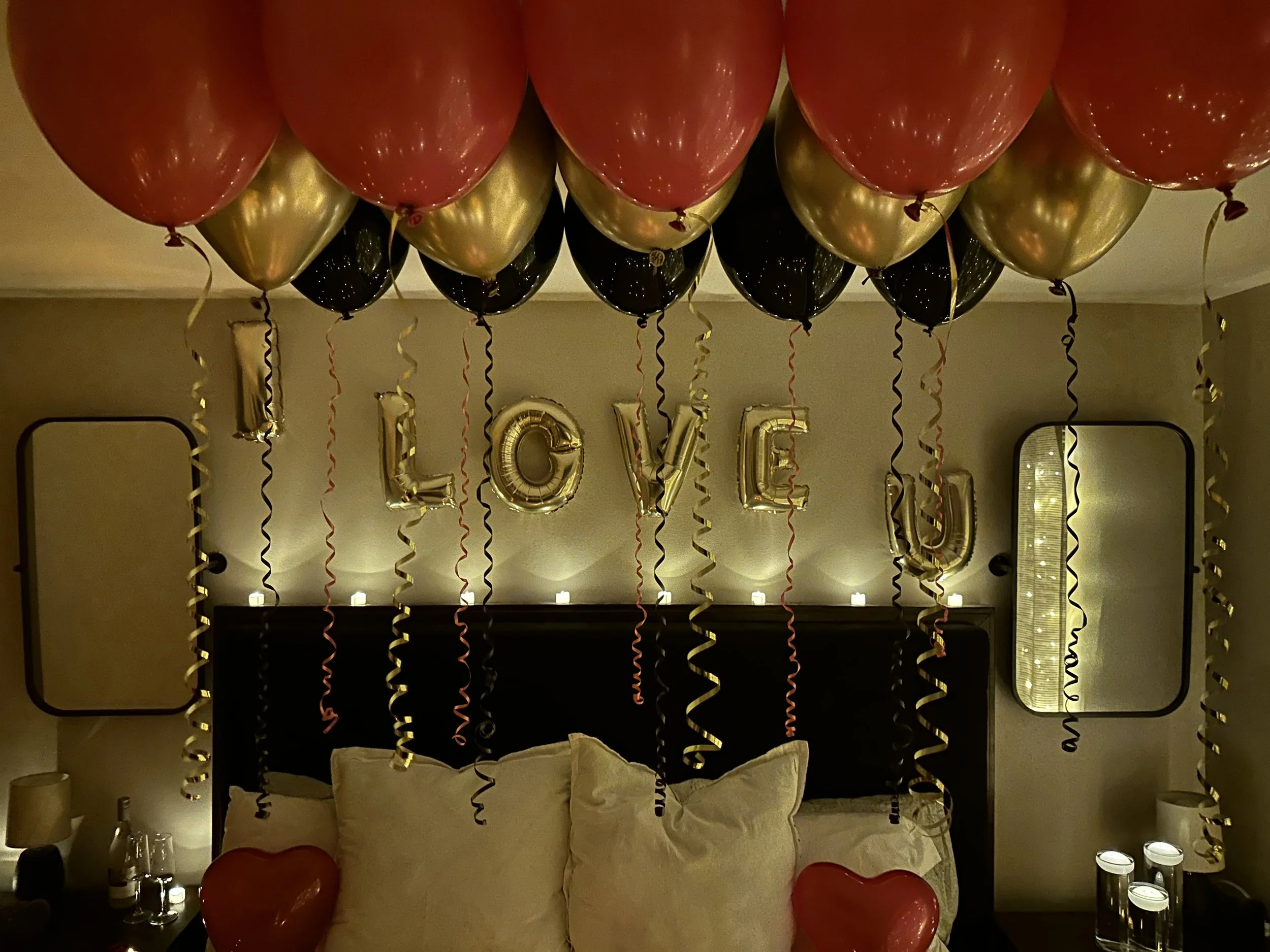 Decorations in a bedroom with a bunch of red, black, and gold balloons, gold letter balloons spelling 'LOVE,' hanging above the bed, with two heart-shaped balloons at the foot of the bed, and candles on bedside tables.