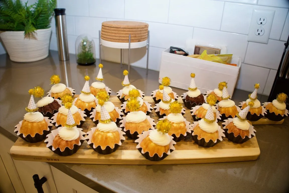 A wooden cutting board with decorated cupcakes, topped with white icing and small party hats with yellow pom-poms, in black and white paper liners, on a kitchen counter.