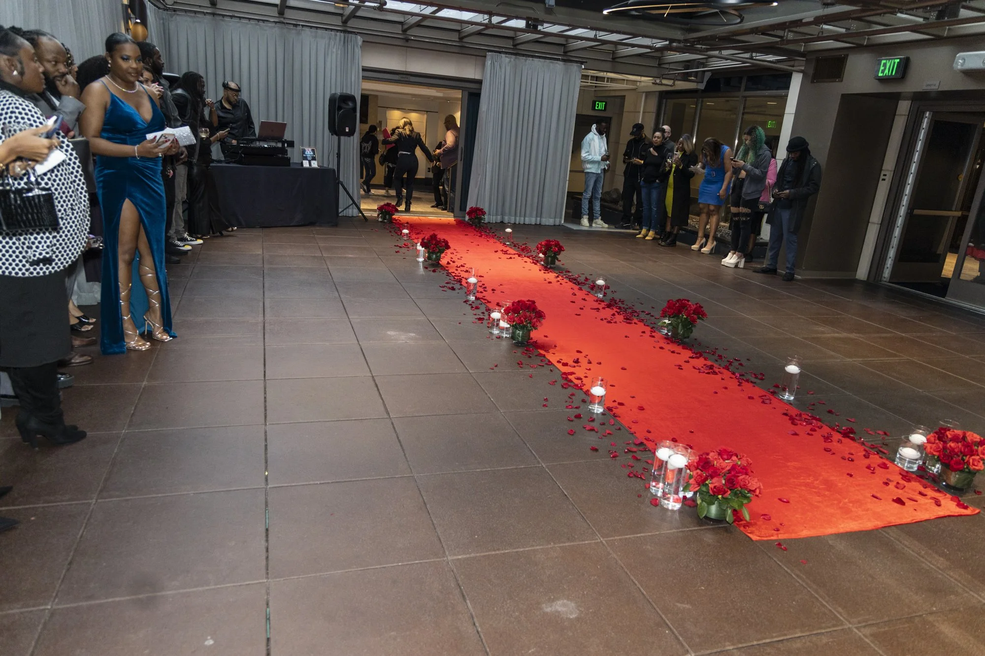A decorated event space with a red carpet lined with red roses and candles, hosting a gathering of people dressed in formal attire indoors.