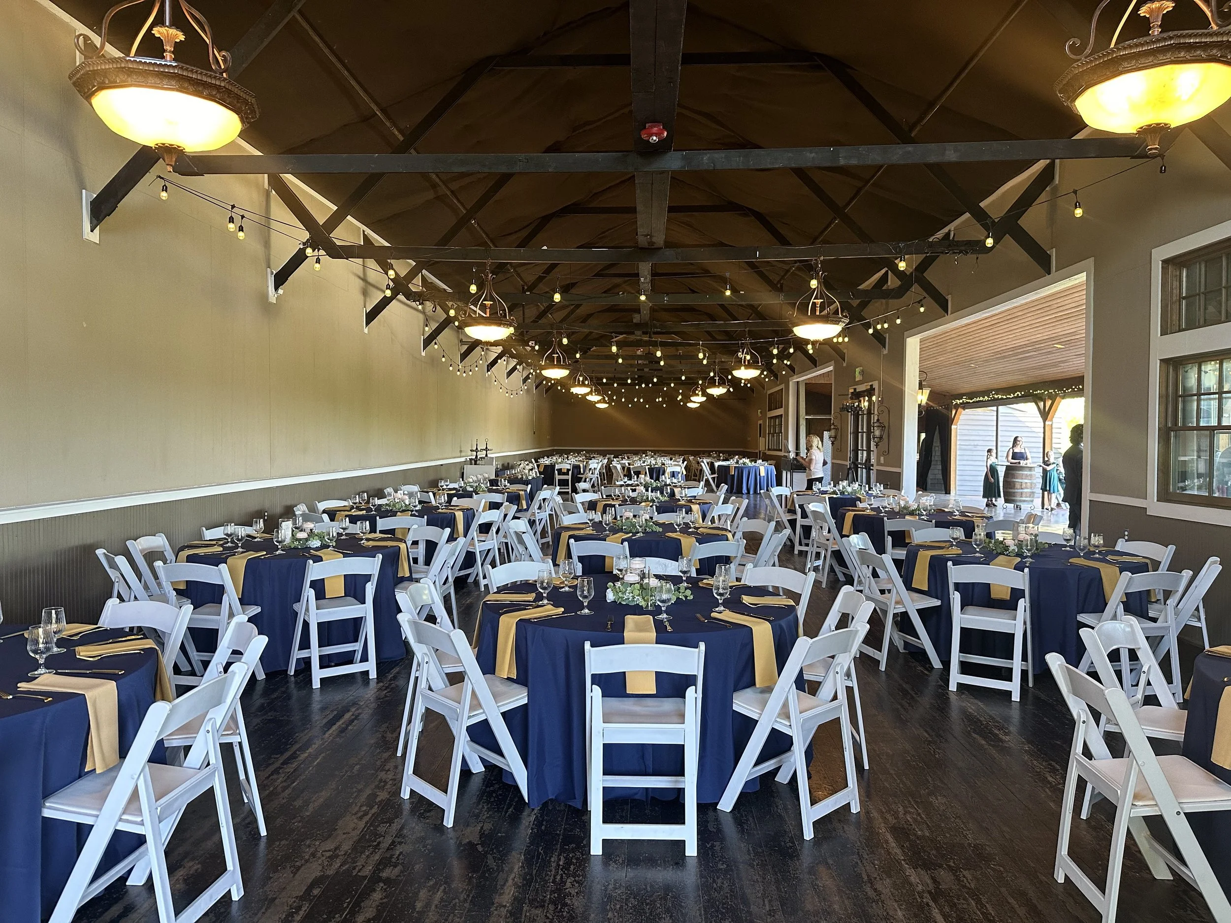 Event hall decorated for a celebration with round tables covered in navy blue tablecloths with gold accents, white chairs, hanging string lights, and ceiling chandeliers; near large windows and outdoor patio area