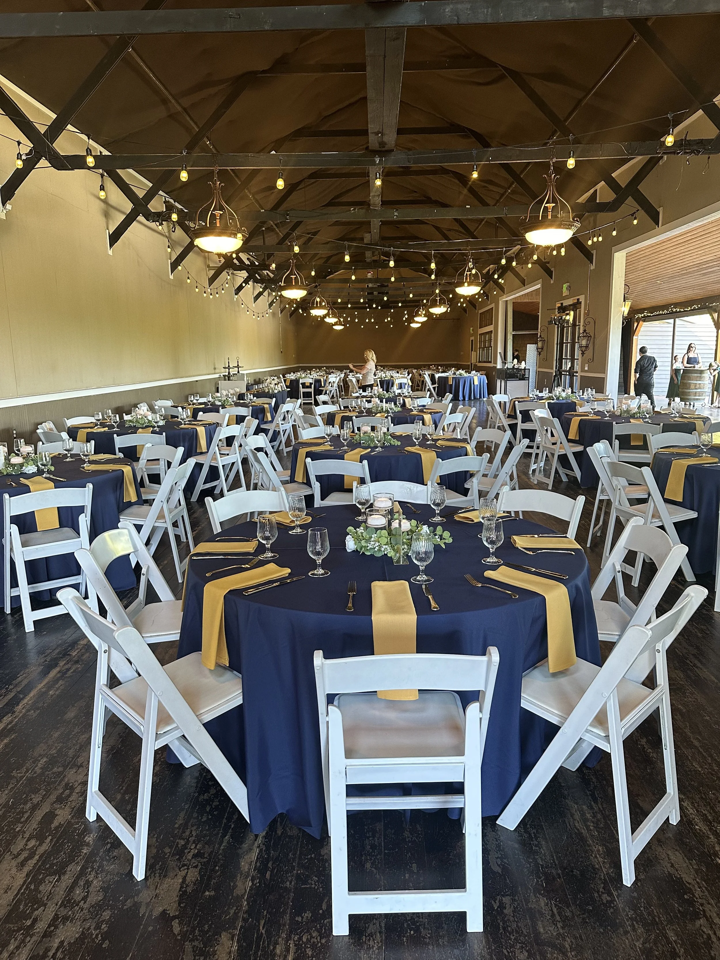 A banquet hall decorated for a formal event with round tables covered in navy blue tablecloths and gold accents, surrounded by white chairs, with hanging lights and a centerpiece with candles and greenery.