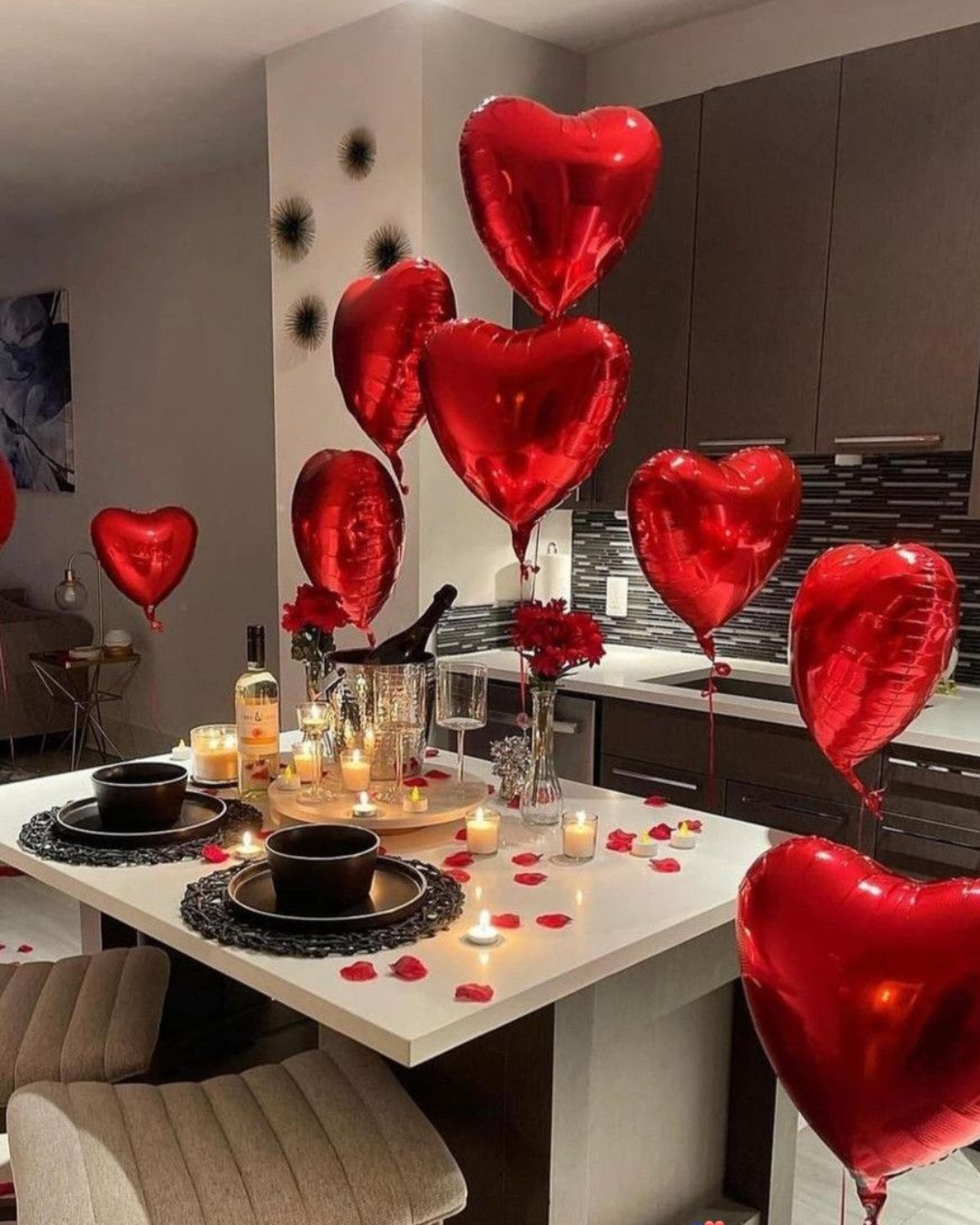 Valentine's Day table setting with red heart-shaped balloons, scattered rose petals, lit candles, and wine bottles in a modern kitchen.
