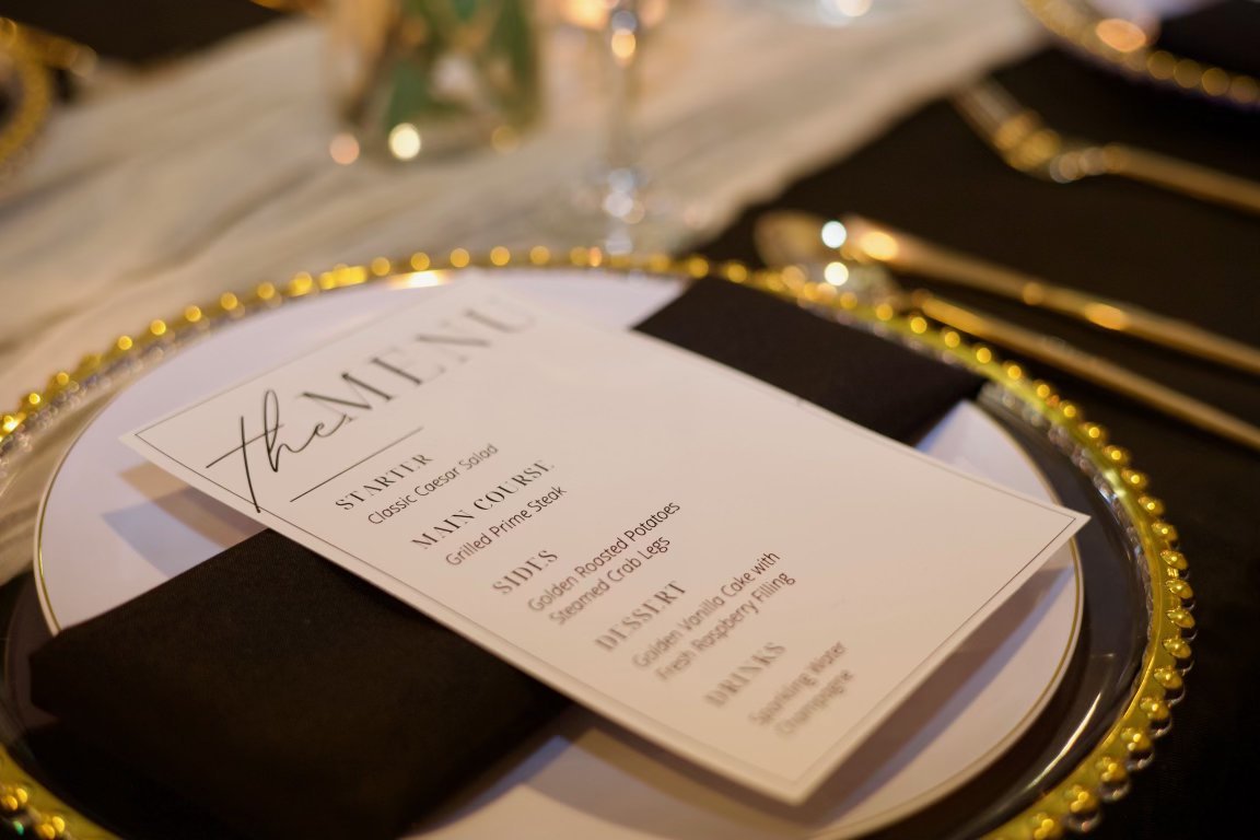 Elegant table setting featuring a decorated plate with a menu, black napkin, and gold beaded rim, suggesting a formal dinner event.