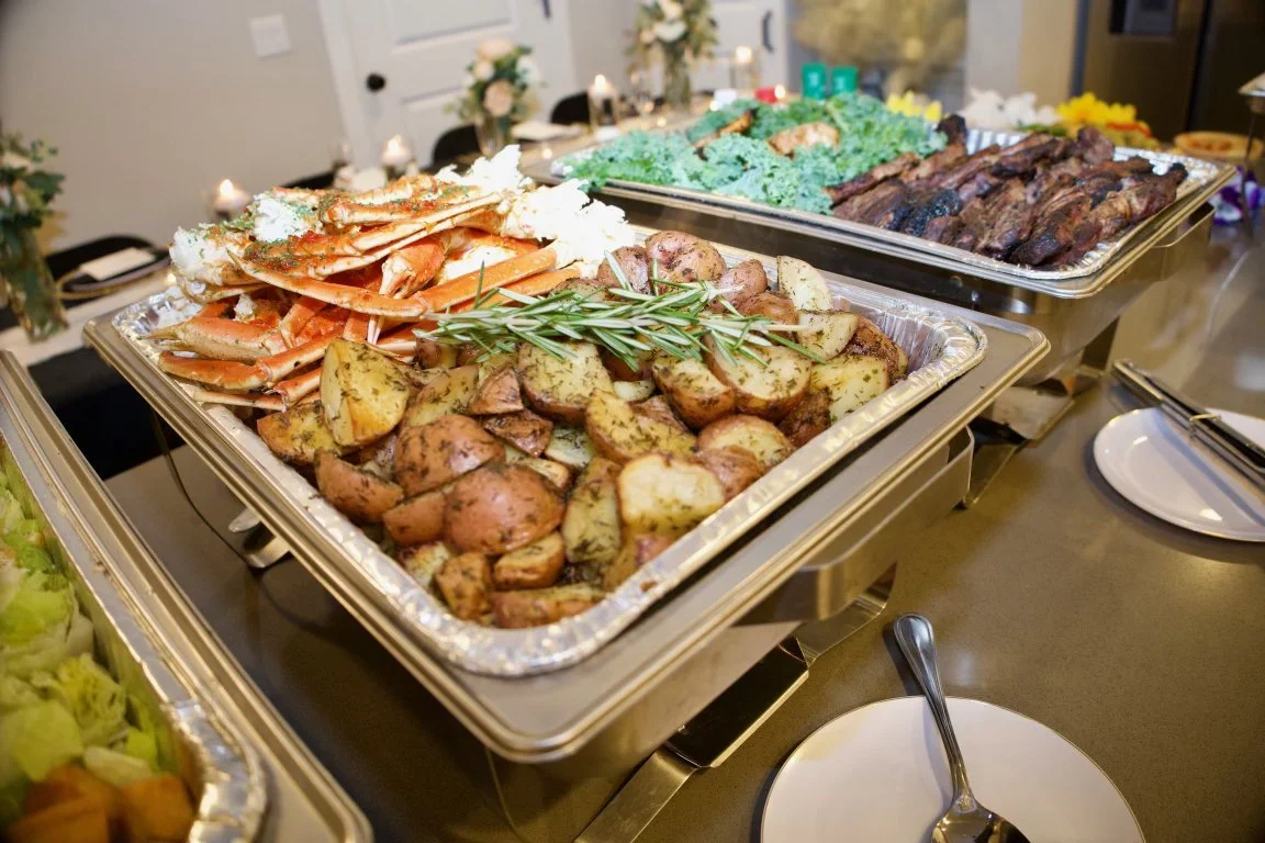 Buffet table with roasted baby potatoes, sprigs of rosemary, crab legs, green vegetable dish, and grilled meat. Decorated with flowers in the background.
