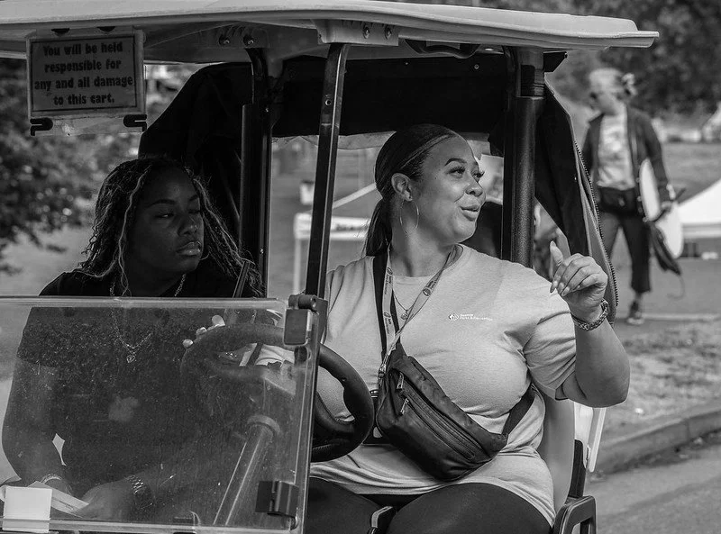 Two women sitting in a golf cart at an outdoor event, with a person walking in the background.