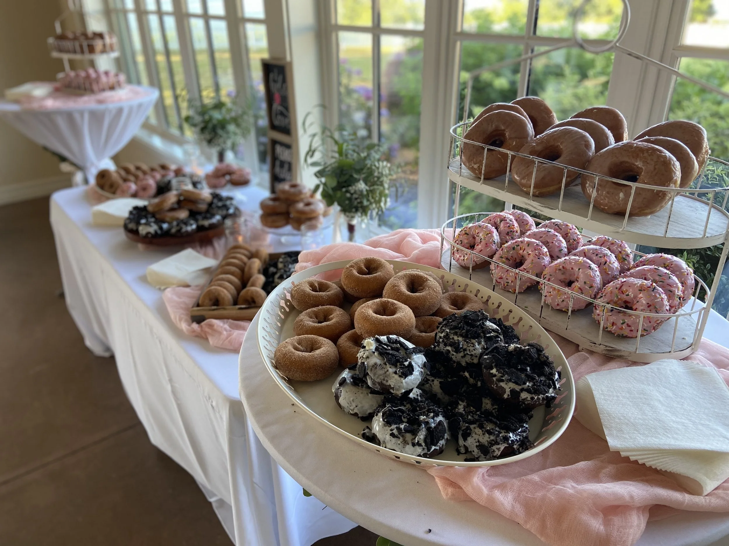 Assorted donuts displayed on a table with pink and white decorations, near a window with greenery outside.