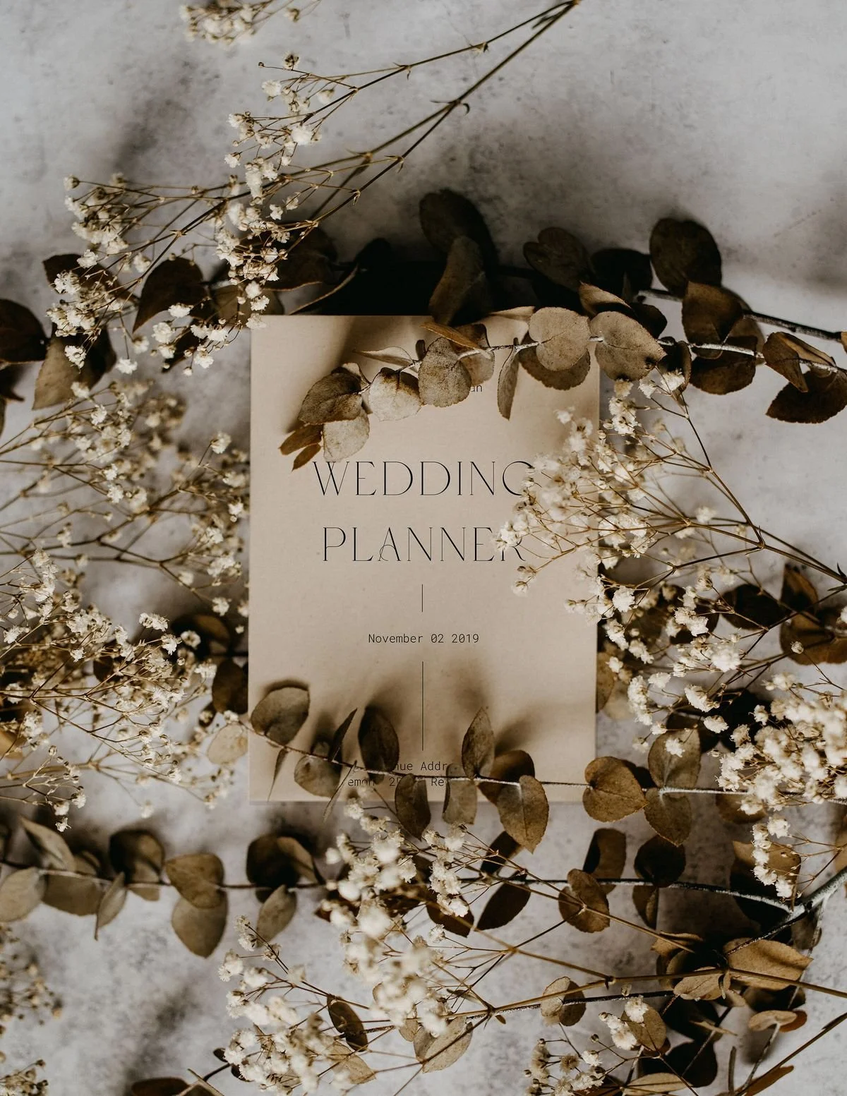 Wedding planner booklet surrounded by dried flowers and leaves on a gray surface.