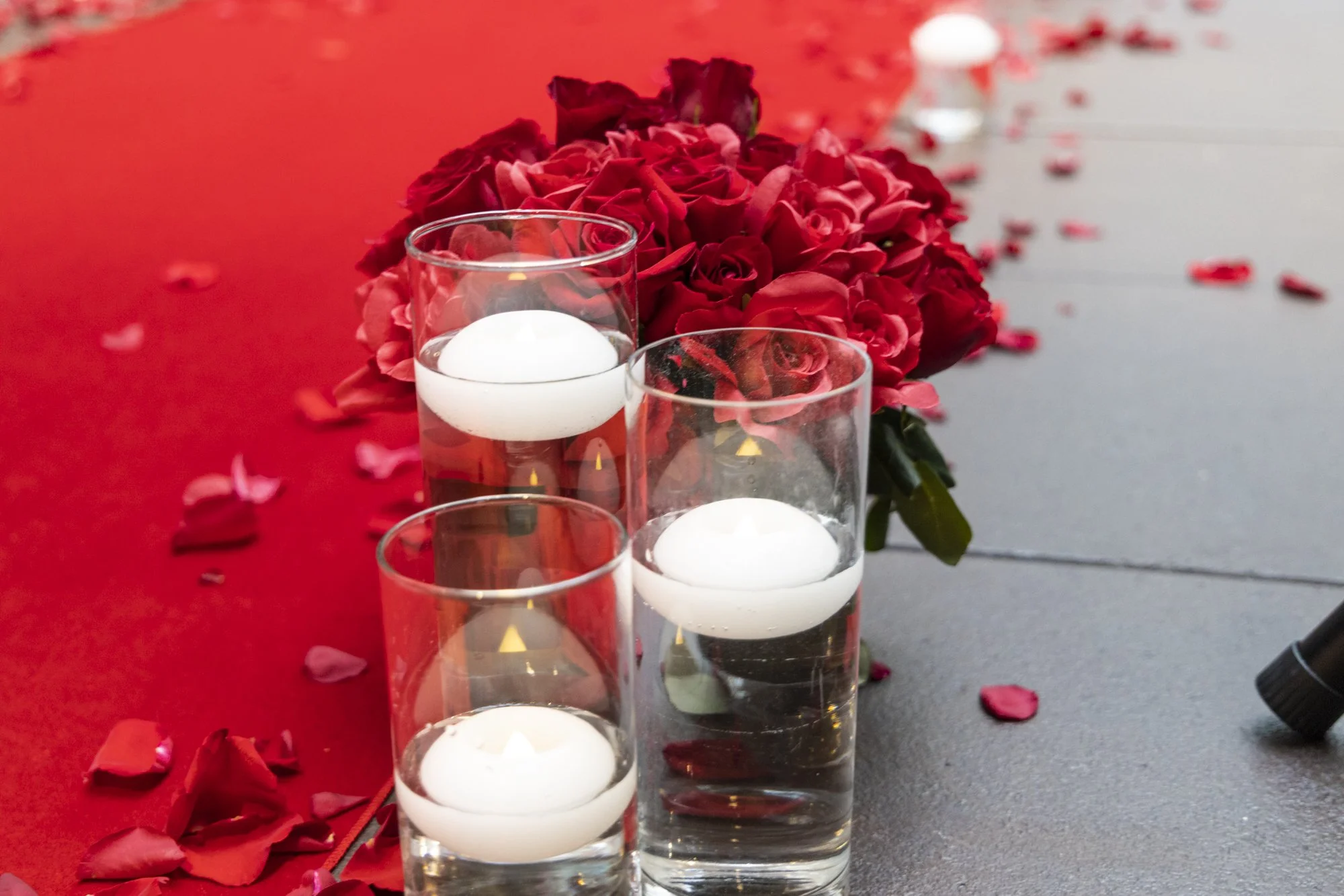 Three glass candles with floating white tealights on a table decorated with red rose petals and a large bouquet of red roses in the background.