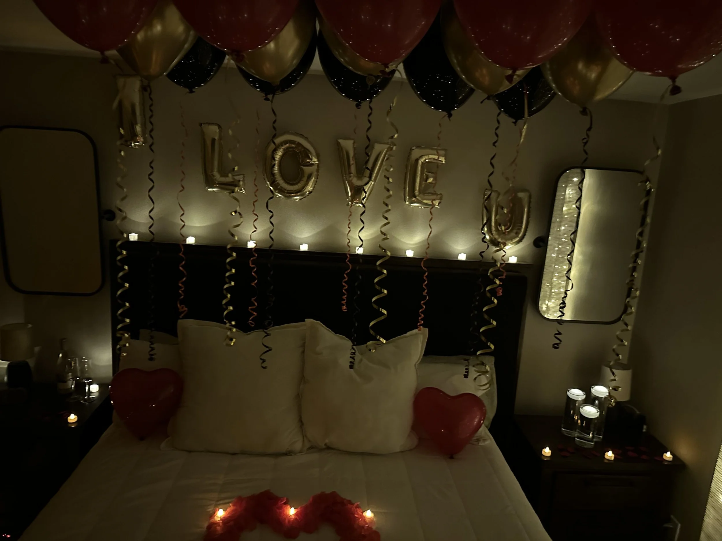 A decorated bedroom with balloons spelling 'LOVE' hanging over the bed, candles, heart-shaped balloons, and rose petals on the bed.