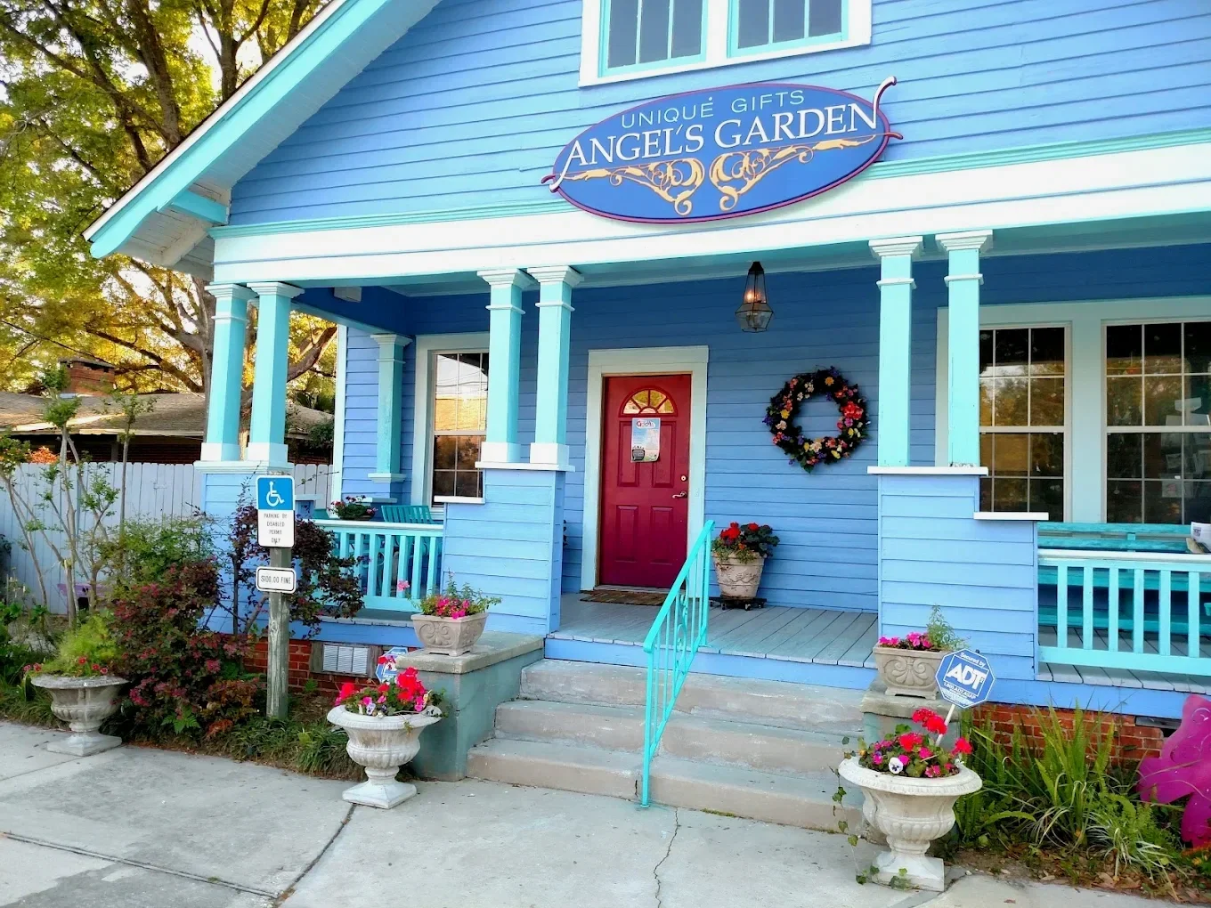 A bright blue storefront with a sign that says 'Unique Gifts – Angel's Garden.' The building has a bright blue siding, a red door, white-framed windows, decorative yellow trim, and potted flowers on the front porch and flower beds.