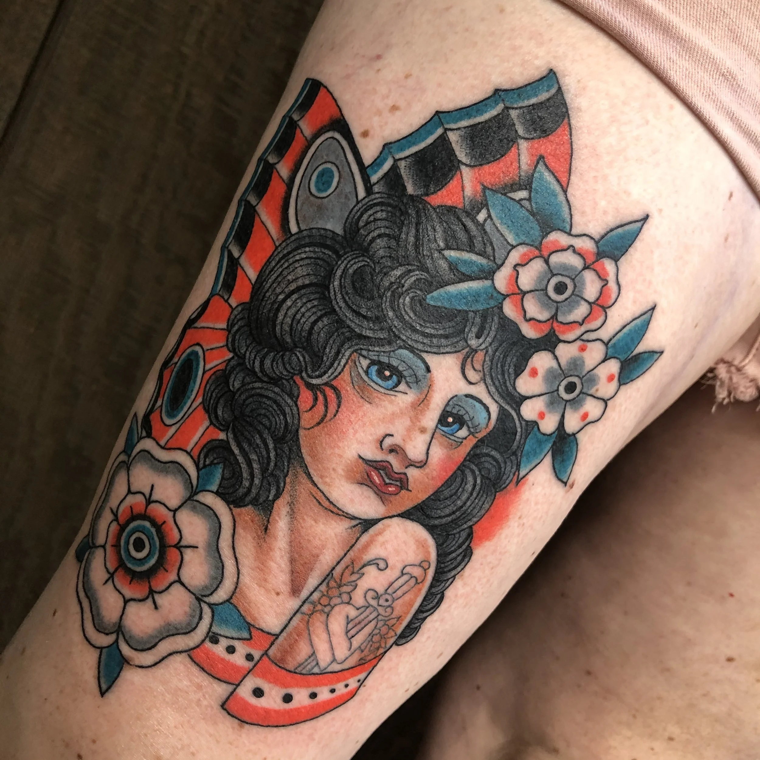Tattoo of a woman with black curly hair, blue eyes, and red lips, surrounded by flowers, with red, black, and blue accents.