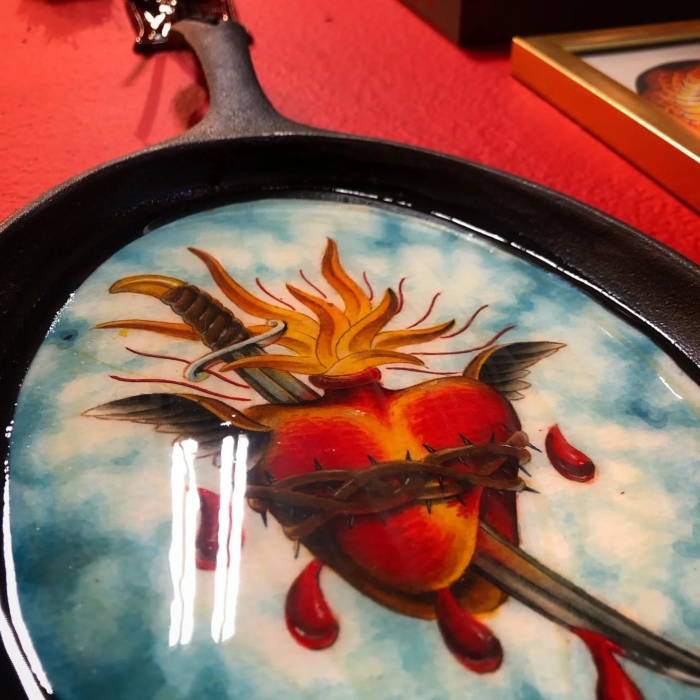 A painted tray with a fiery heart pierced by a sword, surrounded by flames and thorny vine, set against a cloudy sky background.