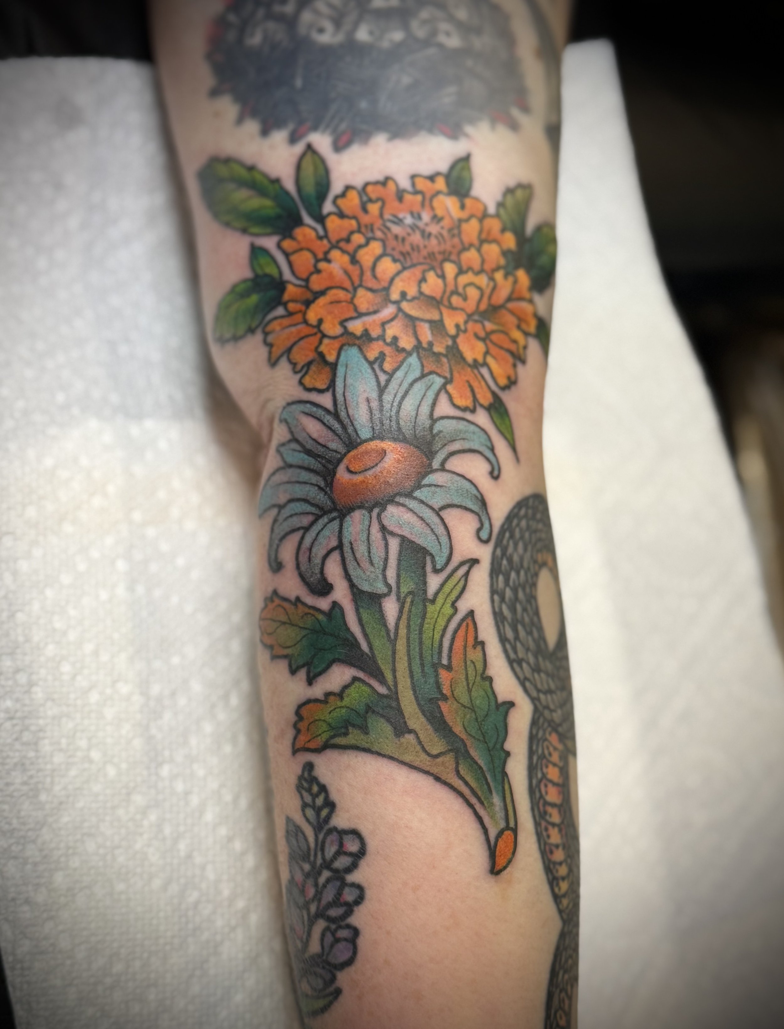 Colorful tattoo of orange flower and white flower with green leaves on person's arm.