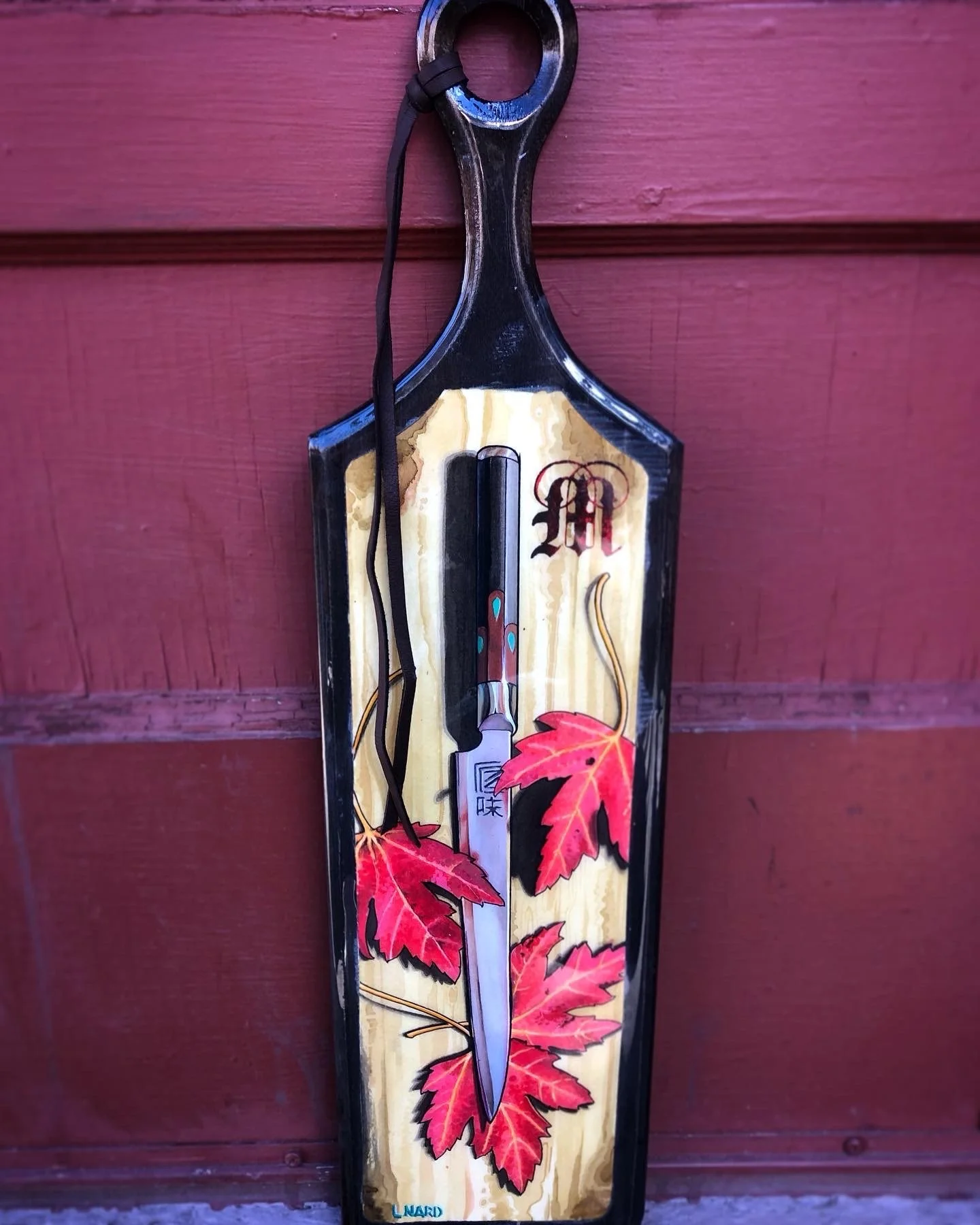 A decorative meat cleaver with a black handle and blade, mounted on wood with red maple leaves design.